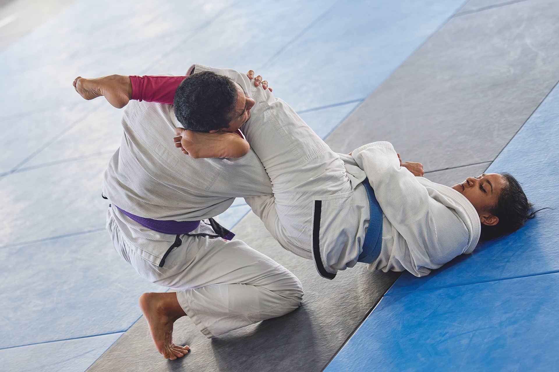 Two martial artists grappling on blue mats, one in a white gi pinning the other.