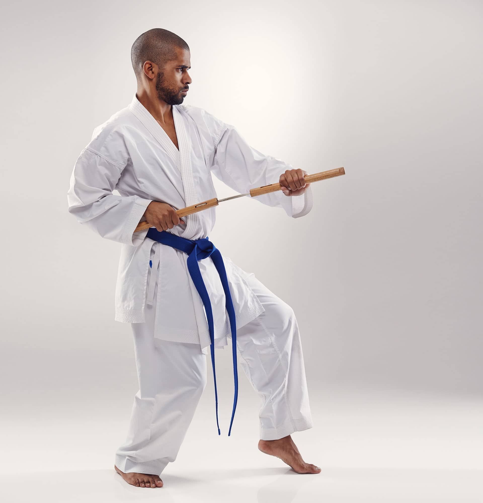 Martial artist in a white gi with blue belt practicing a staff stance on a plain studio background