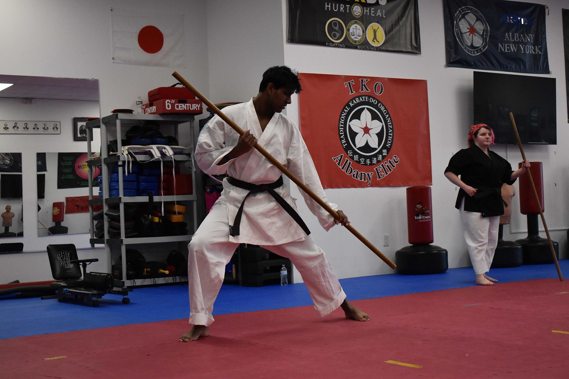 Martial artist in a white gi with blue belt practicing a staff stance on a plain studio background