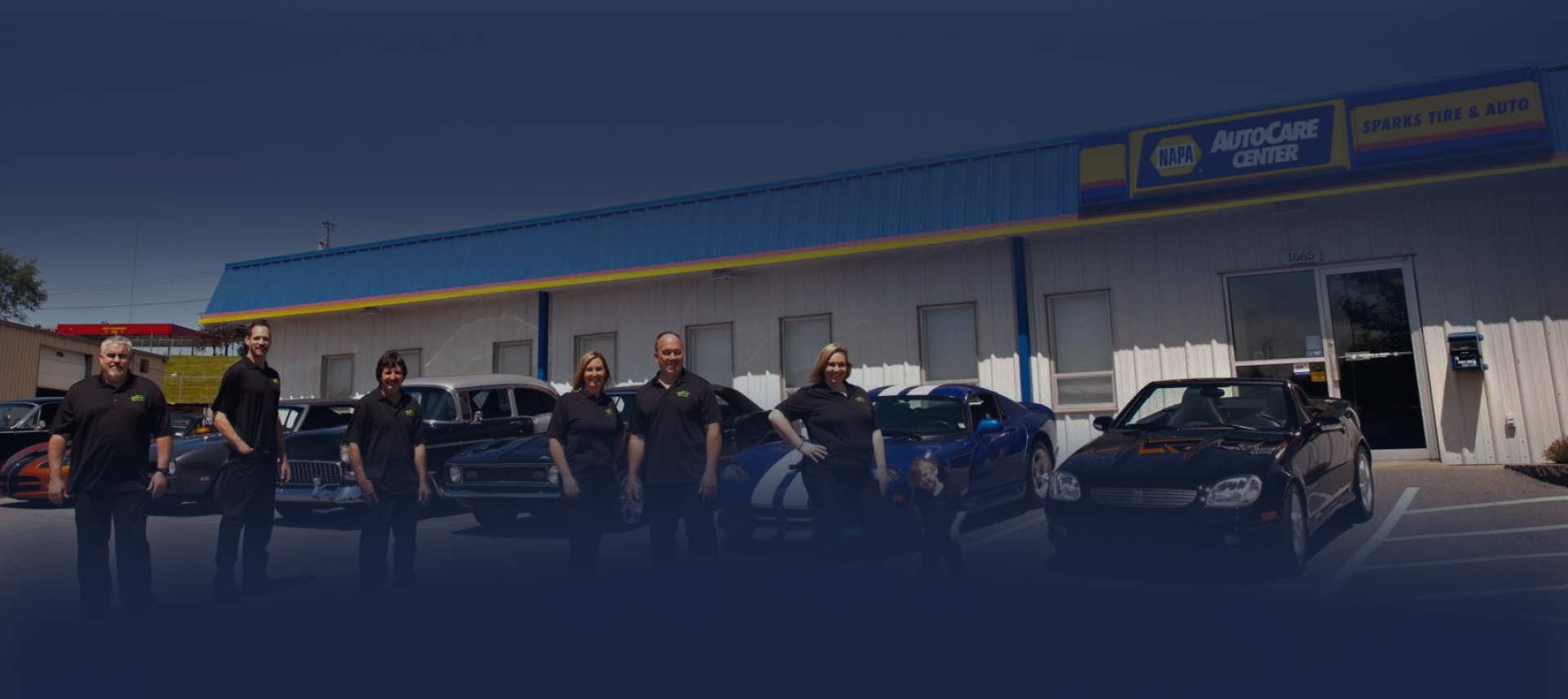 Group of people standing with motorcycles outside a blue auto dealership under a clear sky | Sparks Tire & Auto
