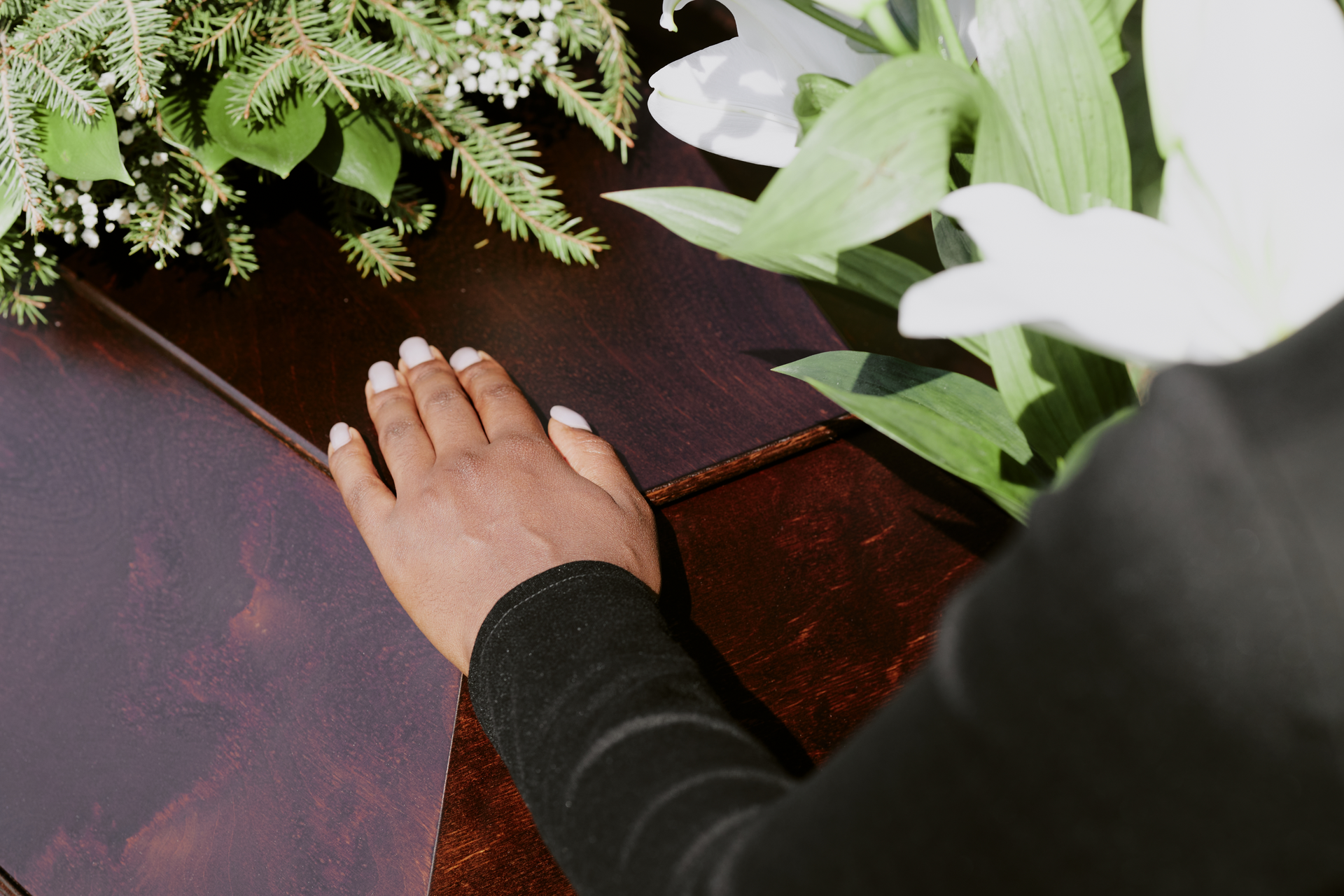 Person's hand on a closed coffin, flowers in background.