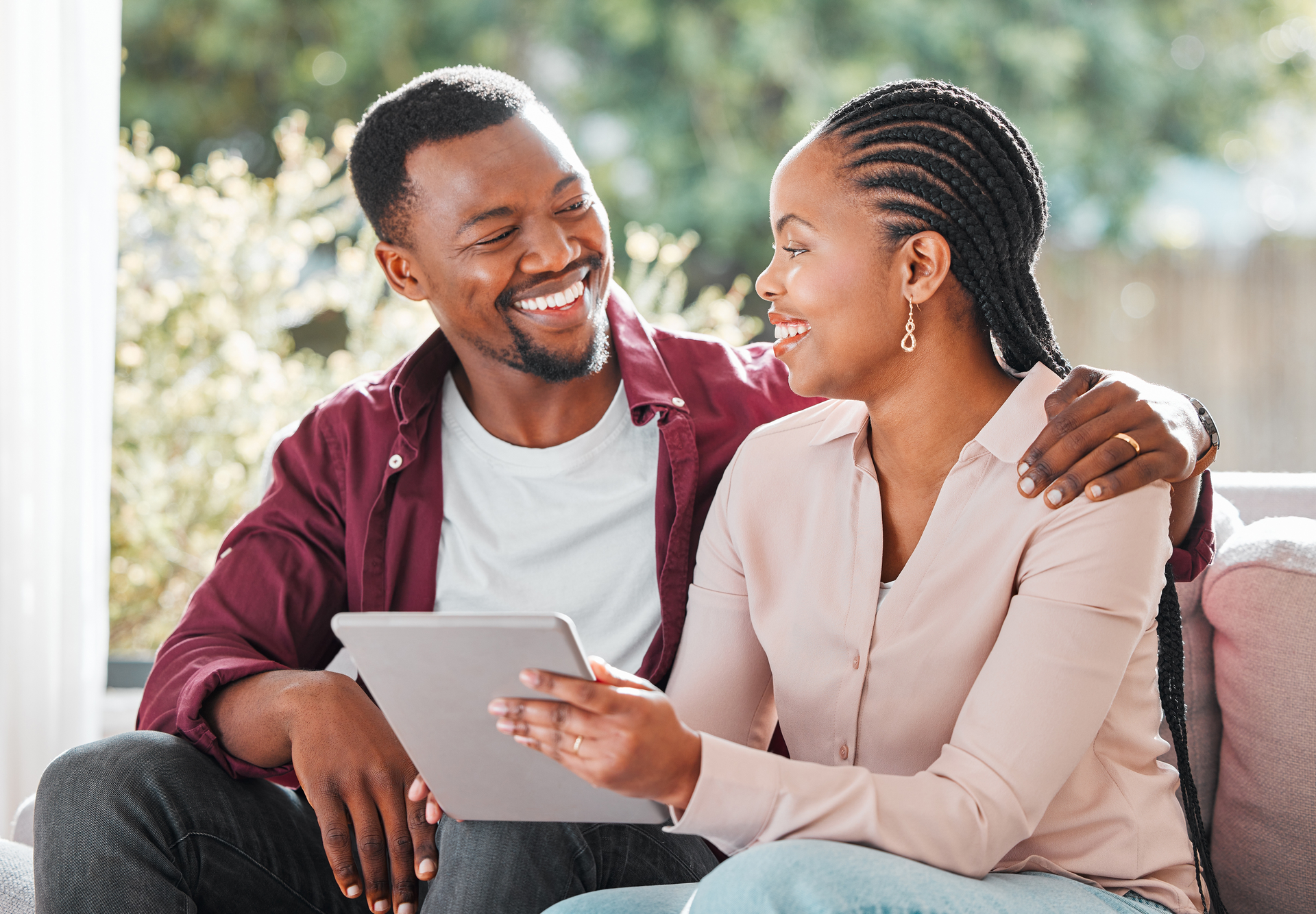 Couple smiling while looking at a tablet on a couch. The man has his arm around the woman. Bright setting.