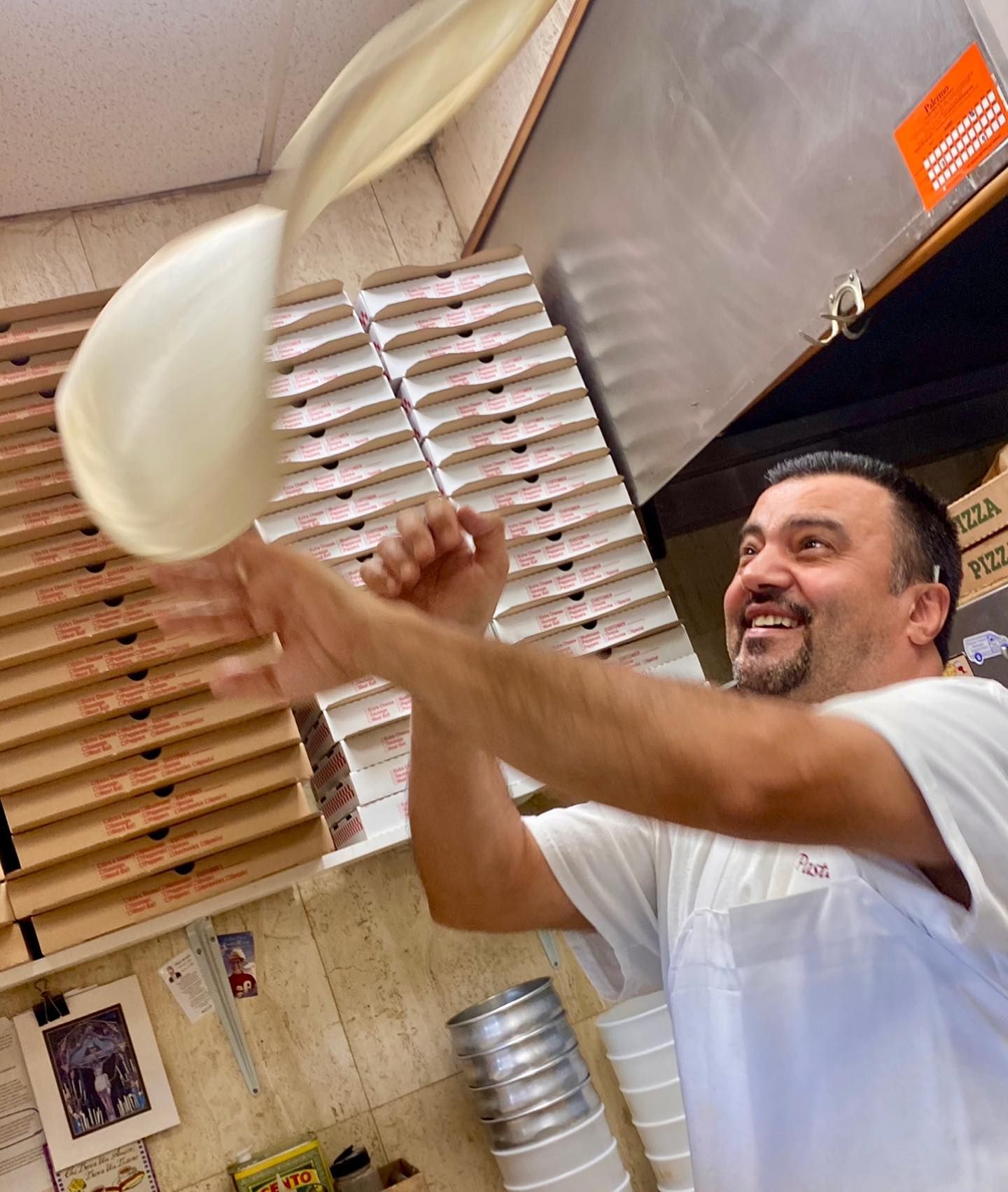 A smiling person tossing pizza dough in a pizzeria. Pizza boxes stacked behind them.