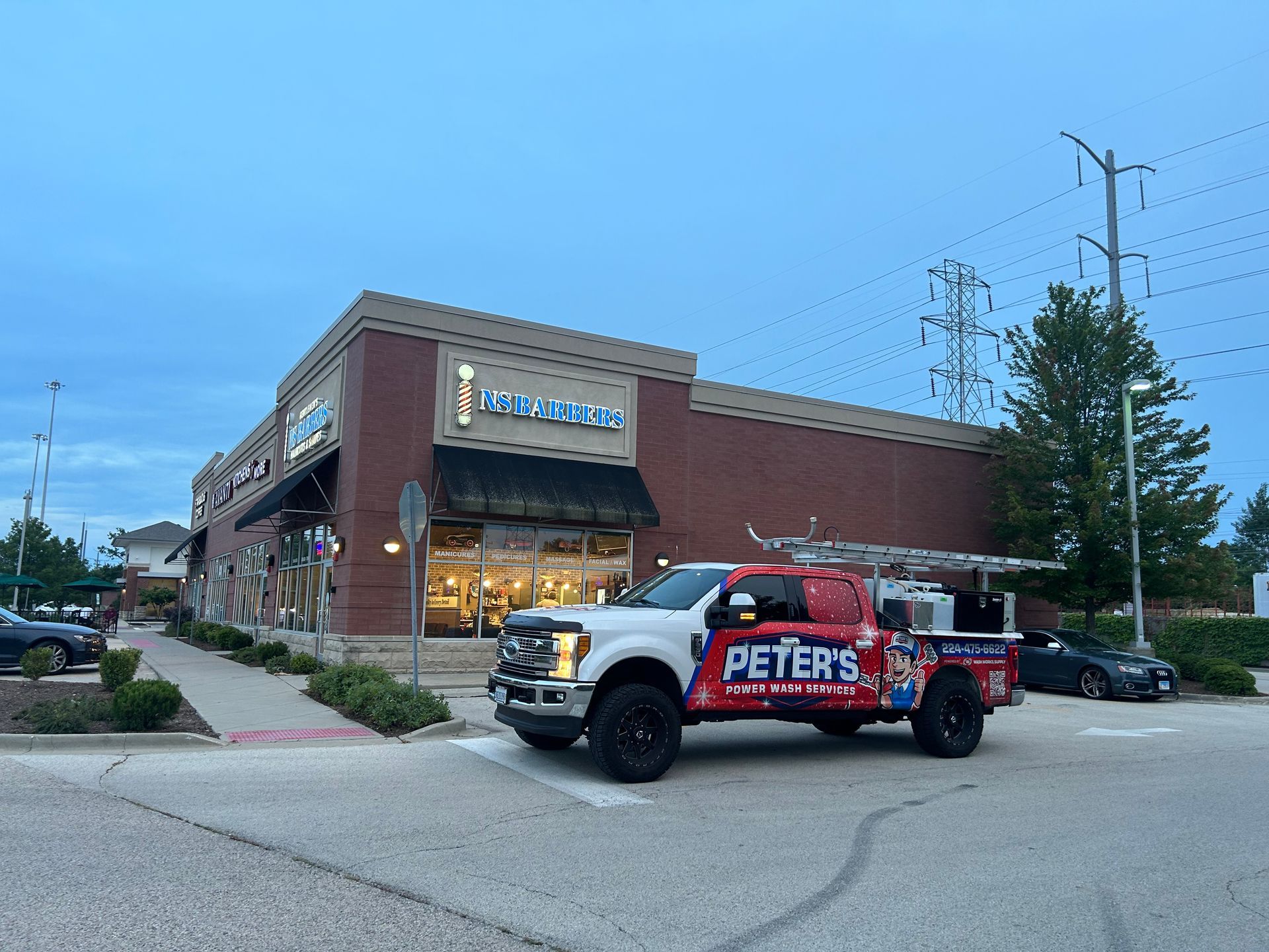 You can see an image of a commercial storefront during a Soft Washing Building Cleaning service, featuring a Peter Power Washing truck parked outside a brick retail building with sidewalks and parking areas freshly cleaned in the early evening.