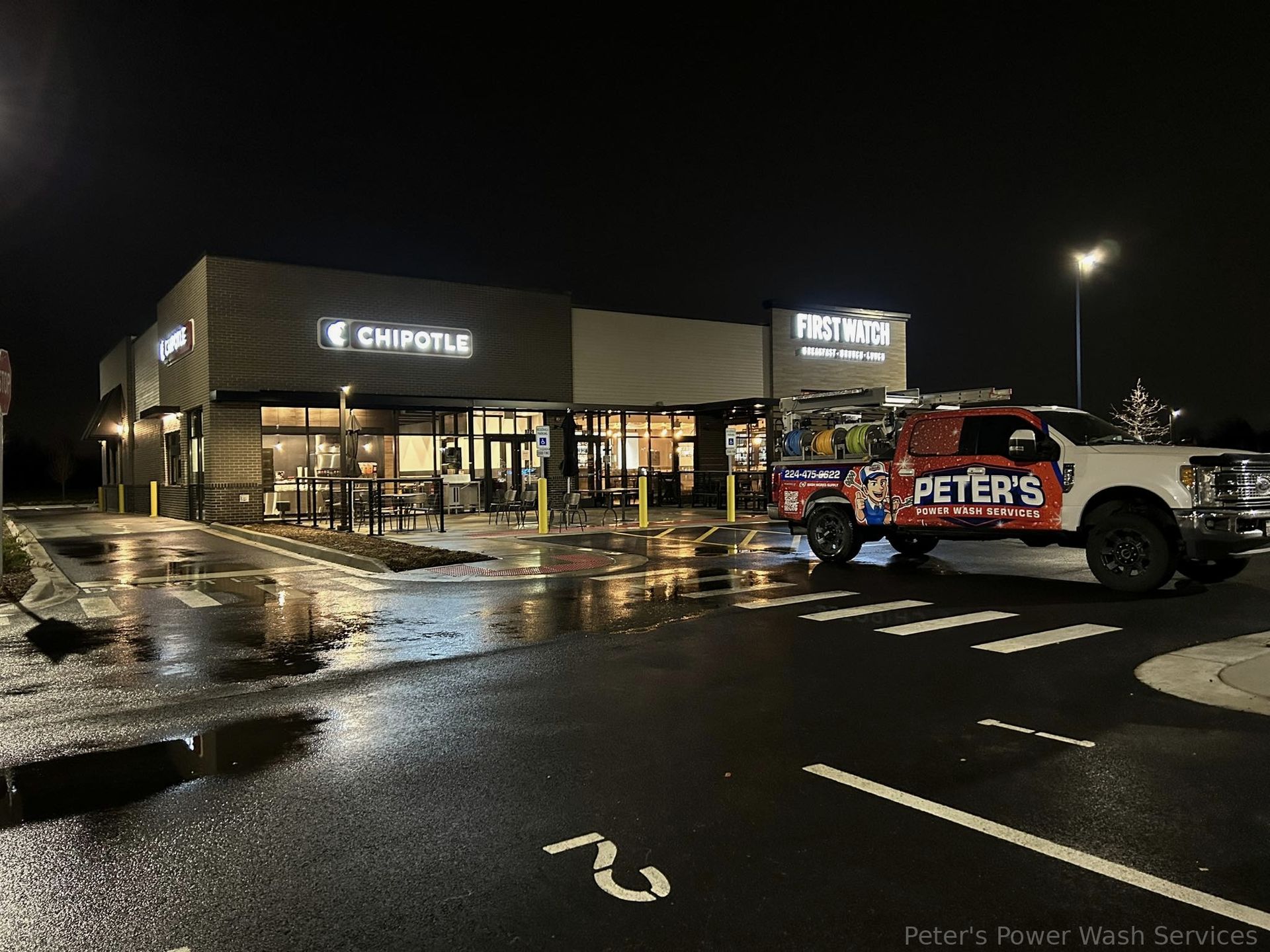 Image of a commercial building at night during a Soft Washing service in Hoffman Estates, featuring a Peter Power Washing truck parked outside a brightly lit storefront with rain-soaked pavement reflecting the building lights and surrounding parking lot.