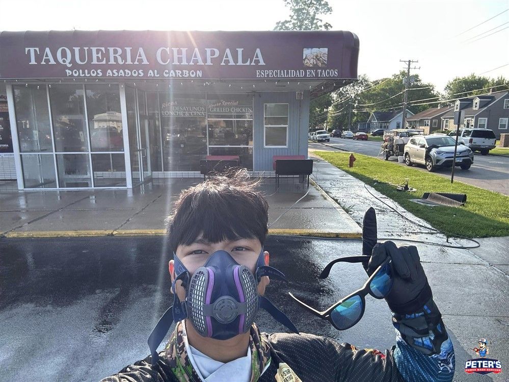 Picture of a worker standing in front of a commercial storefront during a Soft Washing service in Hoffman Estates, wearing protective gear and holding cleaning equipment while the freshly cleaned sidewalk and parking area are visible outside the restaurant building.