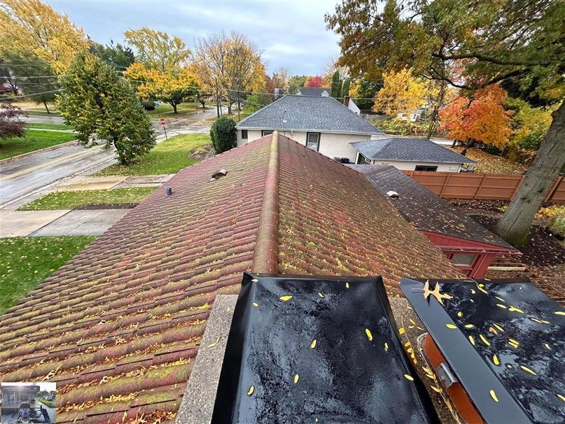 Picture of a residential rooftop during a Soft Washing Roof Washing service, showing a roof covered with leaves and organic buildup being cleaned in a suburban neighborhood with colorful autumn trees surrounding the property.