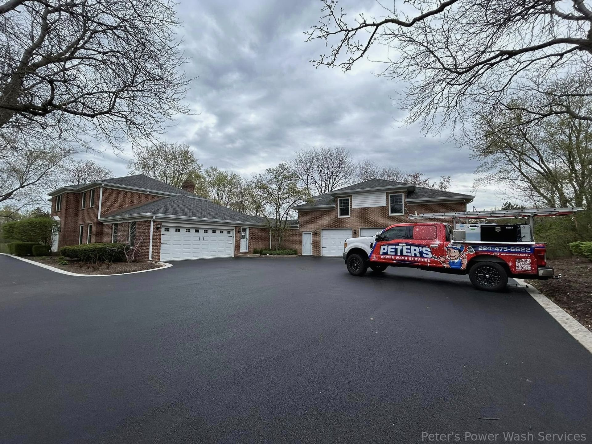 Picture of a freshly cleaned residential driveway during a Soft Washing service in Hoffman Estates, showing a large suburban home with a smooth black driveway surface and a Peter Power Washing truck parked near the front entrance under a cloudy sky.