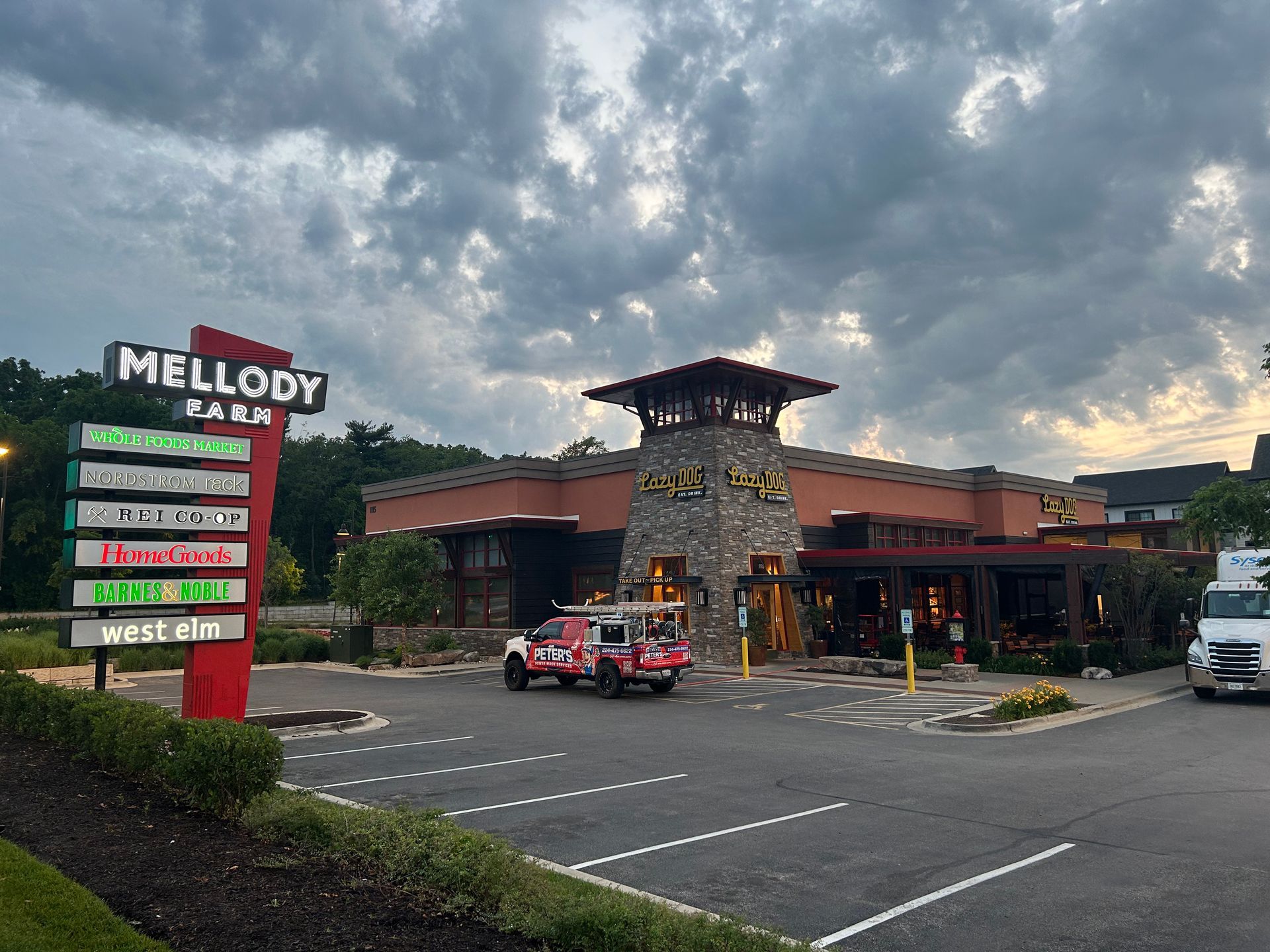 Image of a busy commercial plaza during a Soft Washing Building Cleaning service, showing a restaurant and parking lot area with a Peter Power Washing truck parked near the entrance beneath dramatic cloudy skies.