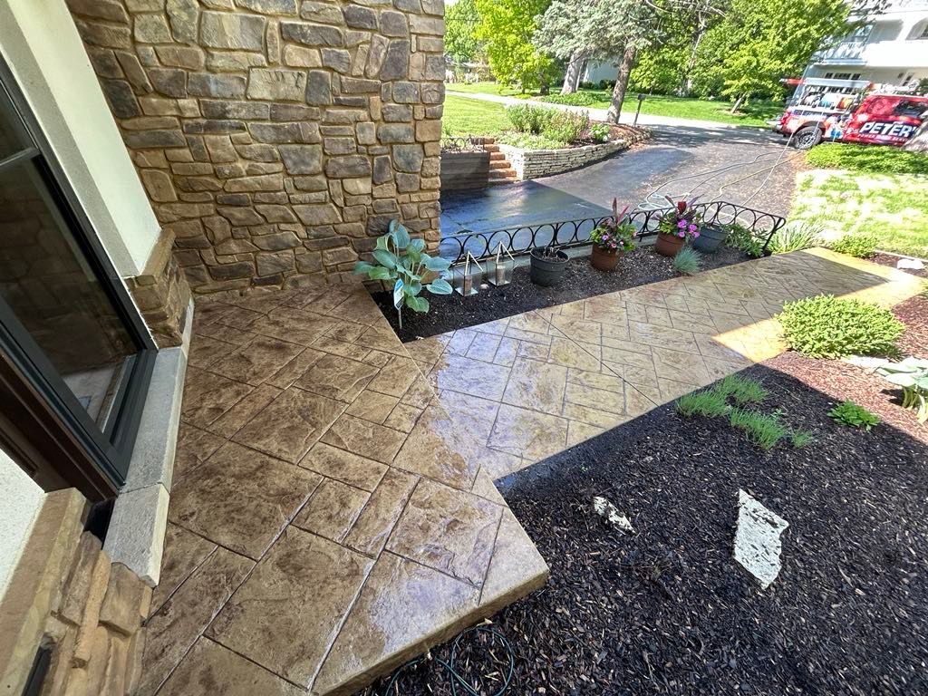 Image of a covered front entry patio during a Soft Washing service in Highland Park, Illinois, featuring freshly cleaned stamped concrete near stone walls, landscaped flower beds, and healthy plants surrounding the walkway.