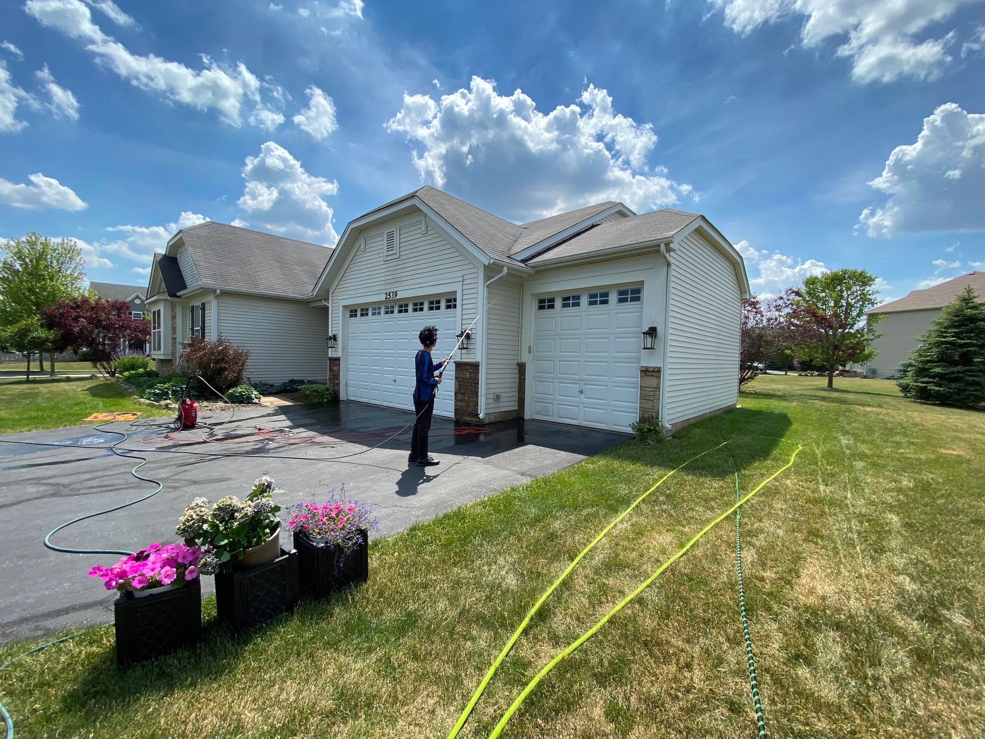 You can see an image of a residential driveway during an HOA Power Washing service, featuring a freshly cleaned concrete surface, landscaped flower beds, and a suburban home under a bright blue sky.
