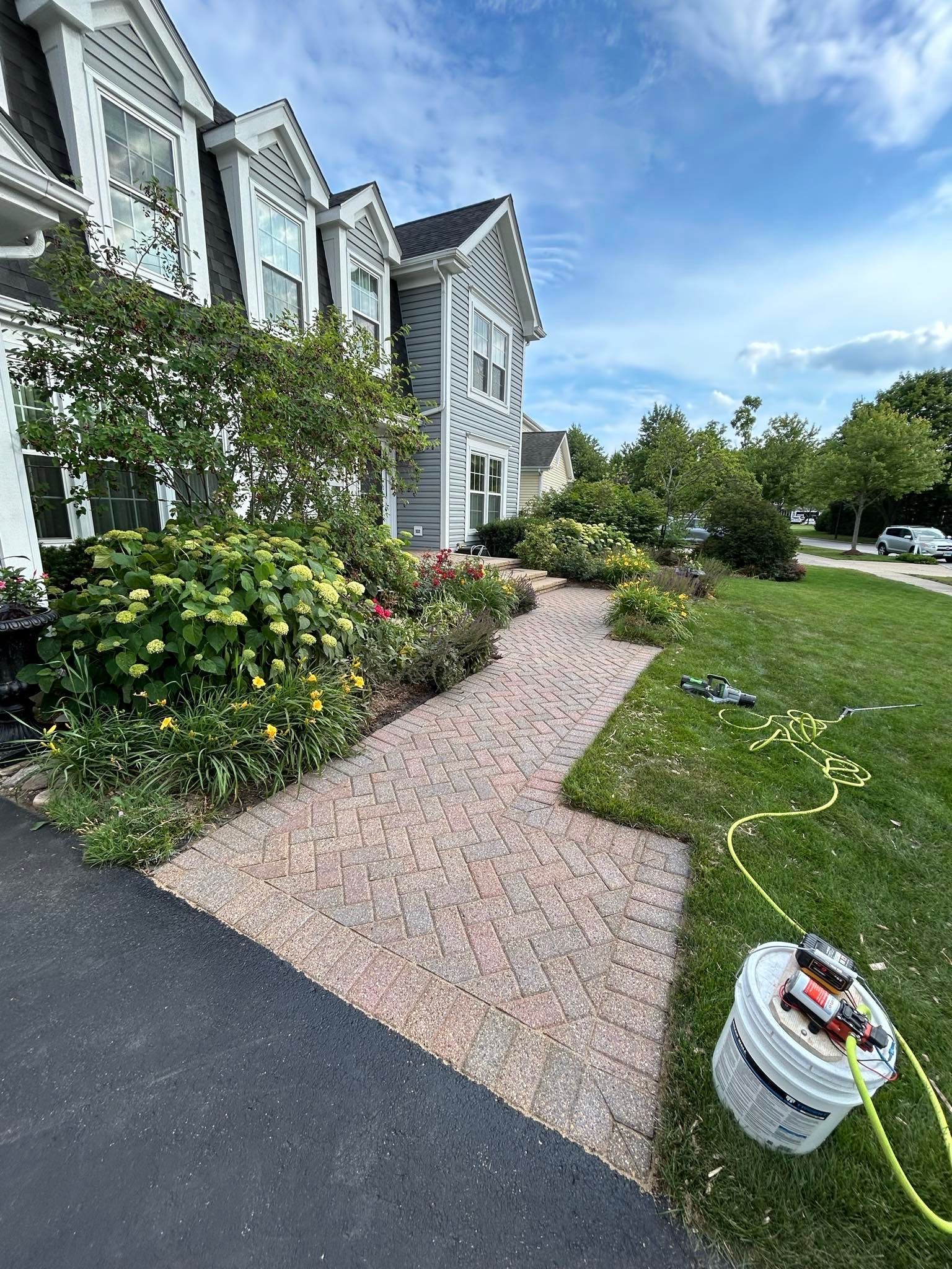 Picture of a large residential home surrounded by landscaping during a Soft Washing service in Highland Park, Illinois, with clean light-gray siding, flowering bushes, and a bright blue sky visible along the front walkway and lawn.