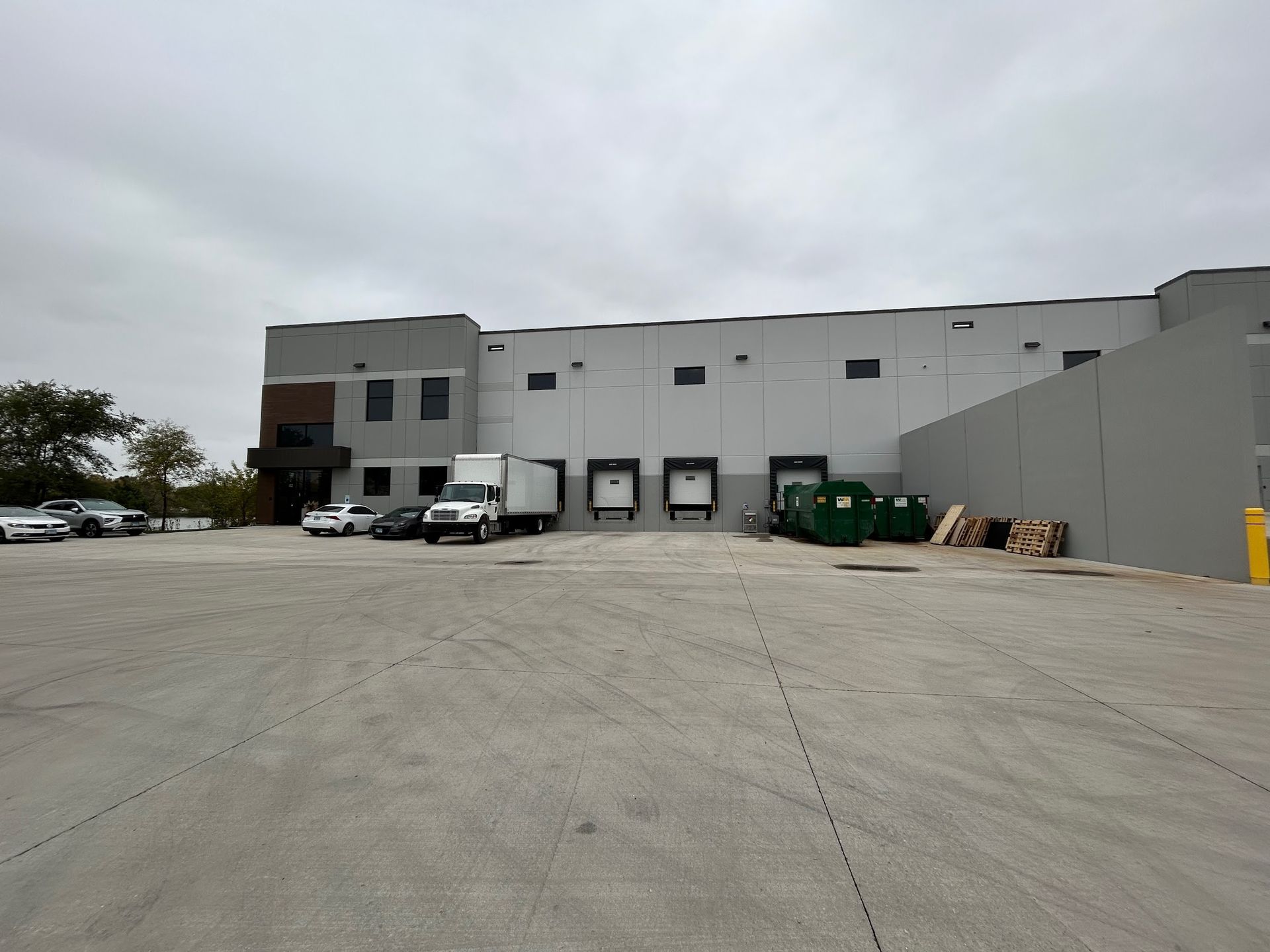 You can see an image of a commercial warehouse property during a Soft Washing Commercial Exterior Cleaning service, featuring a wide clean concrete lot, multiple loading bays, and a large industrial building under an overcast sky.