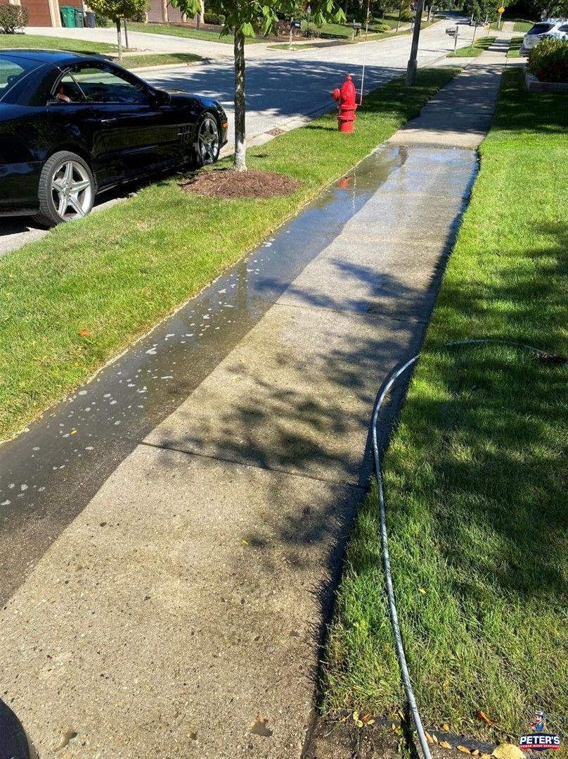 Image of a freshly cleaned residential sidewalk during an HOA Power Washing service, featuring wet concrete walkways beside a parked car, grassy lawn, and neighborhood street.