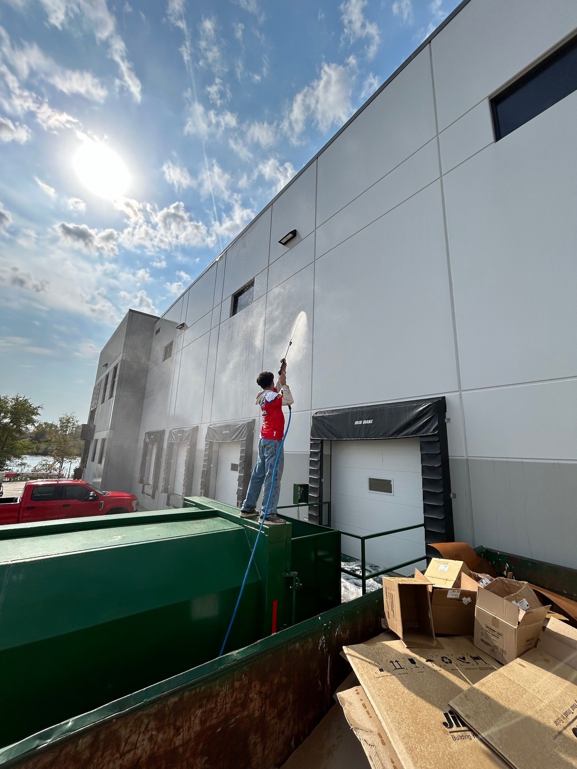 Picture of a worker performing Soft Washing during a Commercial Exterior Cleaning service on a large white industrial building, spraying the exterior wall with cleaning equipment under a bright sky with sunlight and clouds overhead.