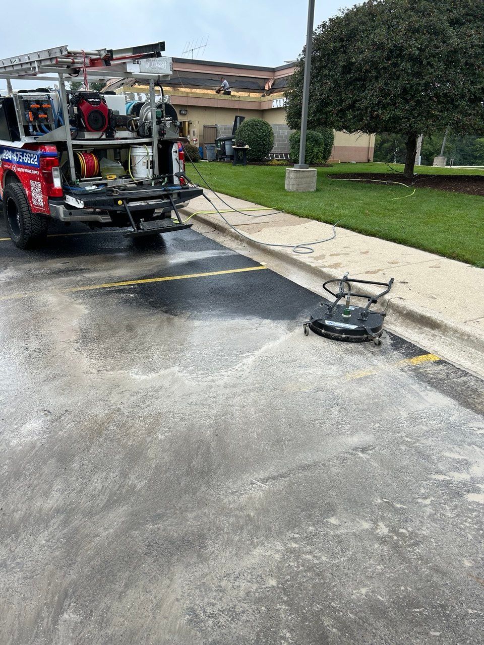 Picture of a Peter Power Washing truck performing a Soft Washing Post Construction Power Wash service in Chicagoland, with surface cleaning equipment washing a commercial sidewalk and parking area beside a landscaped building entrance.