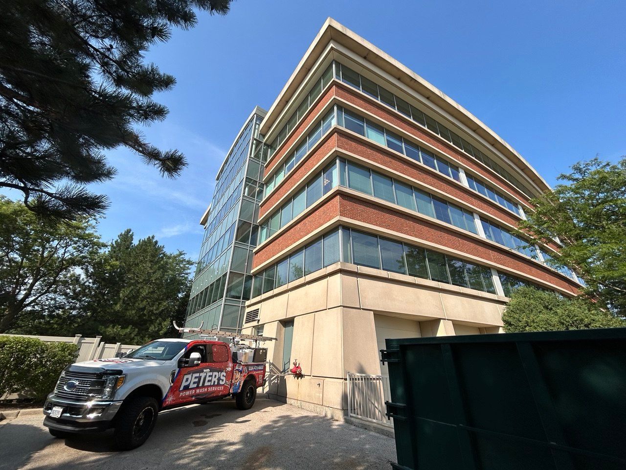 Picture of a modern multi-story office building with large glass windows and orange panel accents under a clear blue sky, viewed from street level beside a Peter Power Washing service van during a Commercial Concrete Cleaning project.