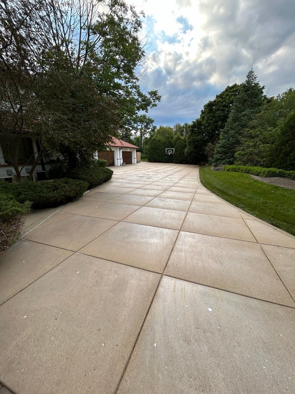 You can see an image of a long residential driveway during a Soft Washing Concrete Sealing service, featuring a freshly cleaned and sealed concrete surface surrounded by trees, landscaping, and a large suburban property under a cloudy evening sky.