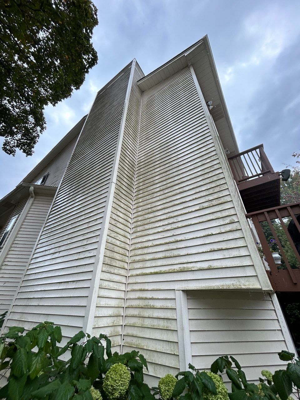Image of the exterior siding of a tall residential home during a Soft Washing House Washing service, captured from a low angle to show the freshly cleaned siding, upper balcony, and cloudy sky above the house.