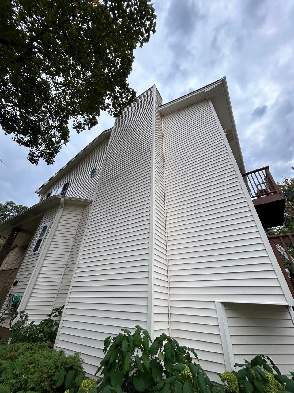 You can see an image of the side exterior of a multi-story residential home during a Soft Washing House Washing service, featuring tall freshly cleaned siding, elevated wooden decks, and surrounding trees viewed from ground level against an overcast sky.