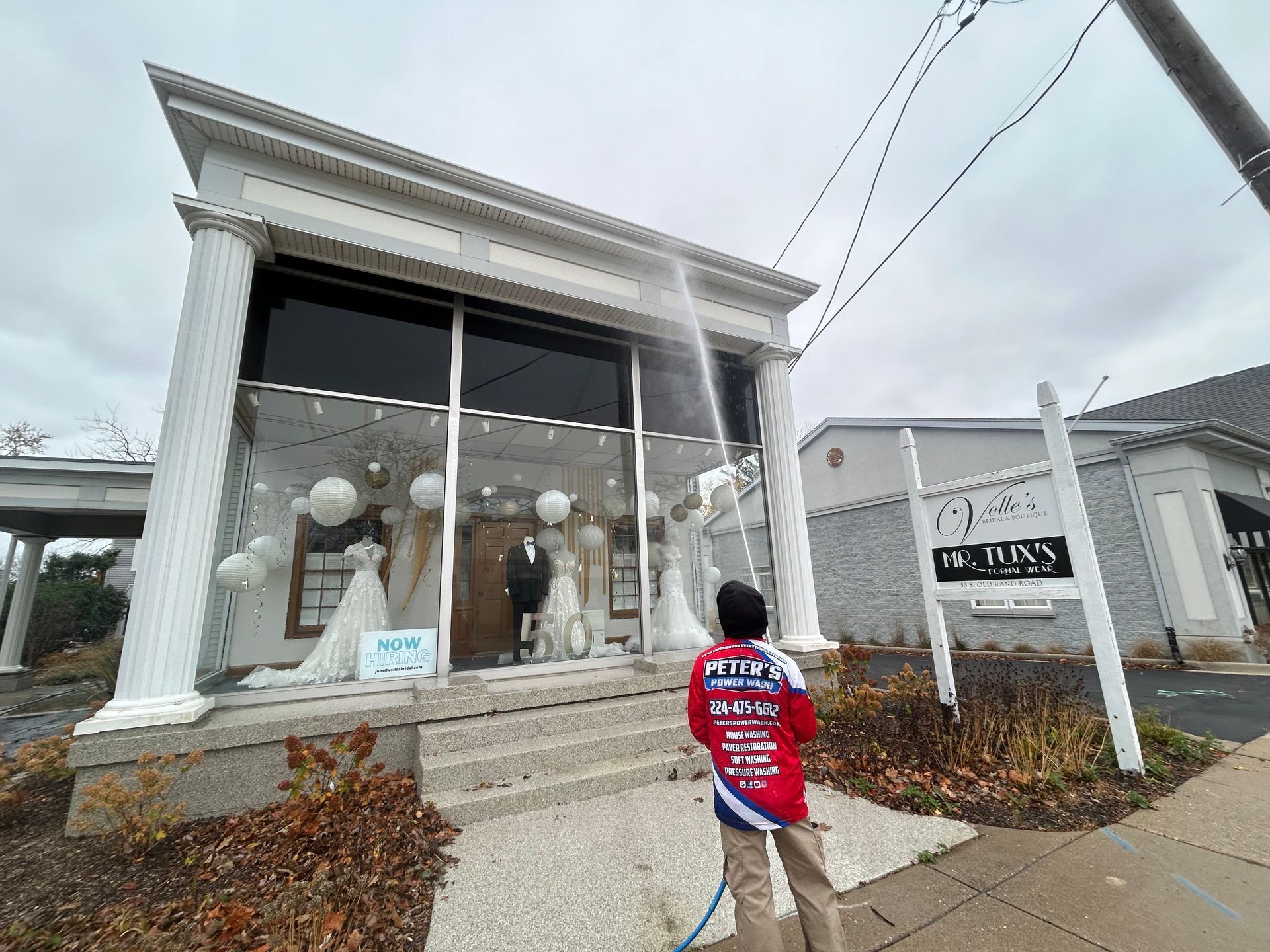 Picture of a Peter Power Washing technician performing a Soft Washing power washing service for walls and windows outside a modern commercial storefront, spraying large glass windows and white exterior walls while standing on the sidewalk under a cloudy sky.