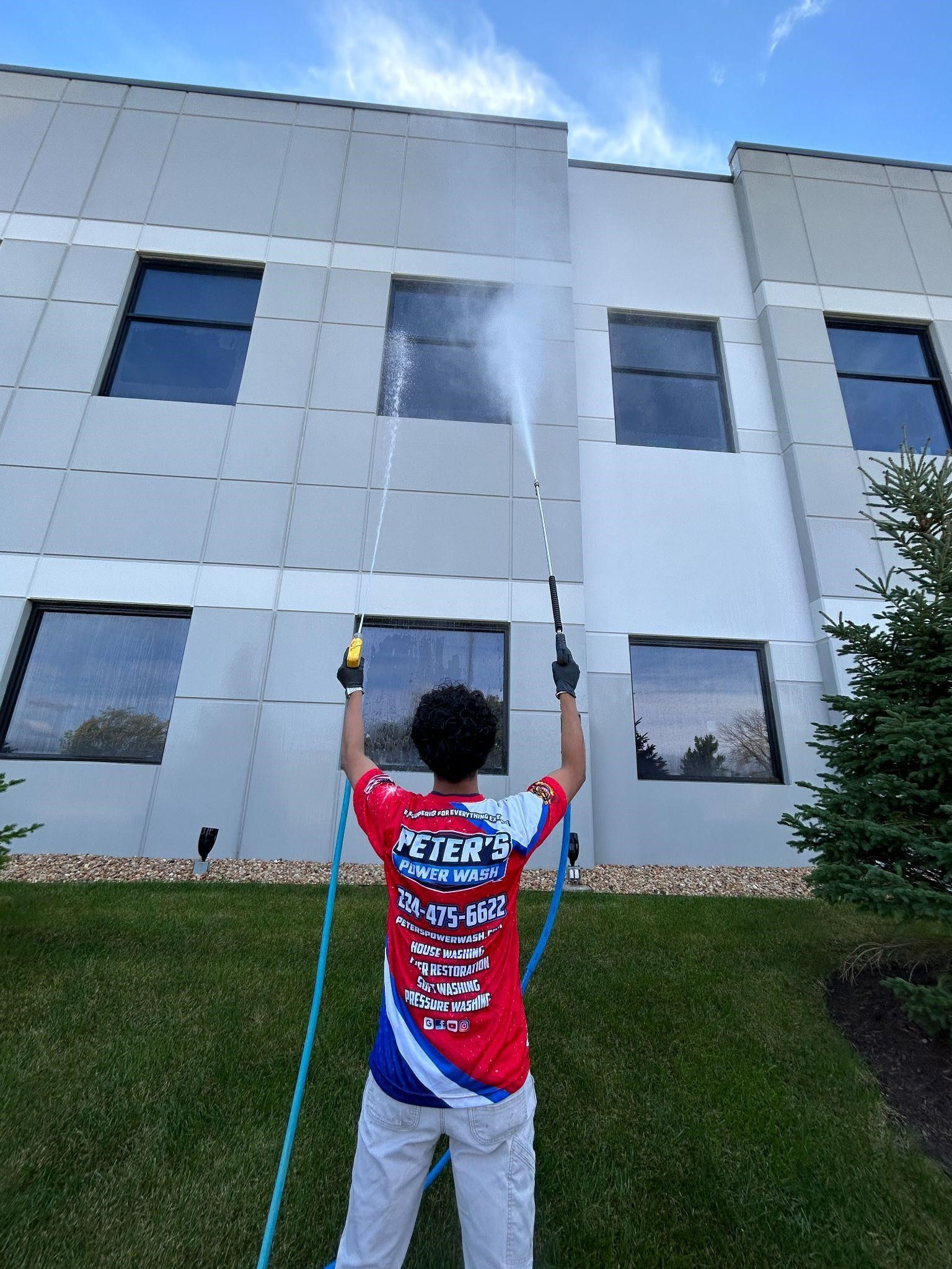 Picture of a commercial building exterior during a Soft Washing Building Cleaning service in Chicagoland, showing water being sprayed onto tall white exterior walls to remove dirt and buildup under a partly cloudy sky.