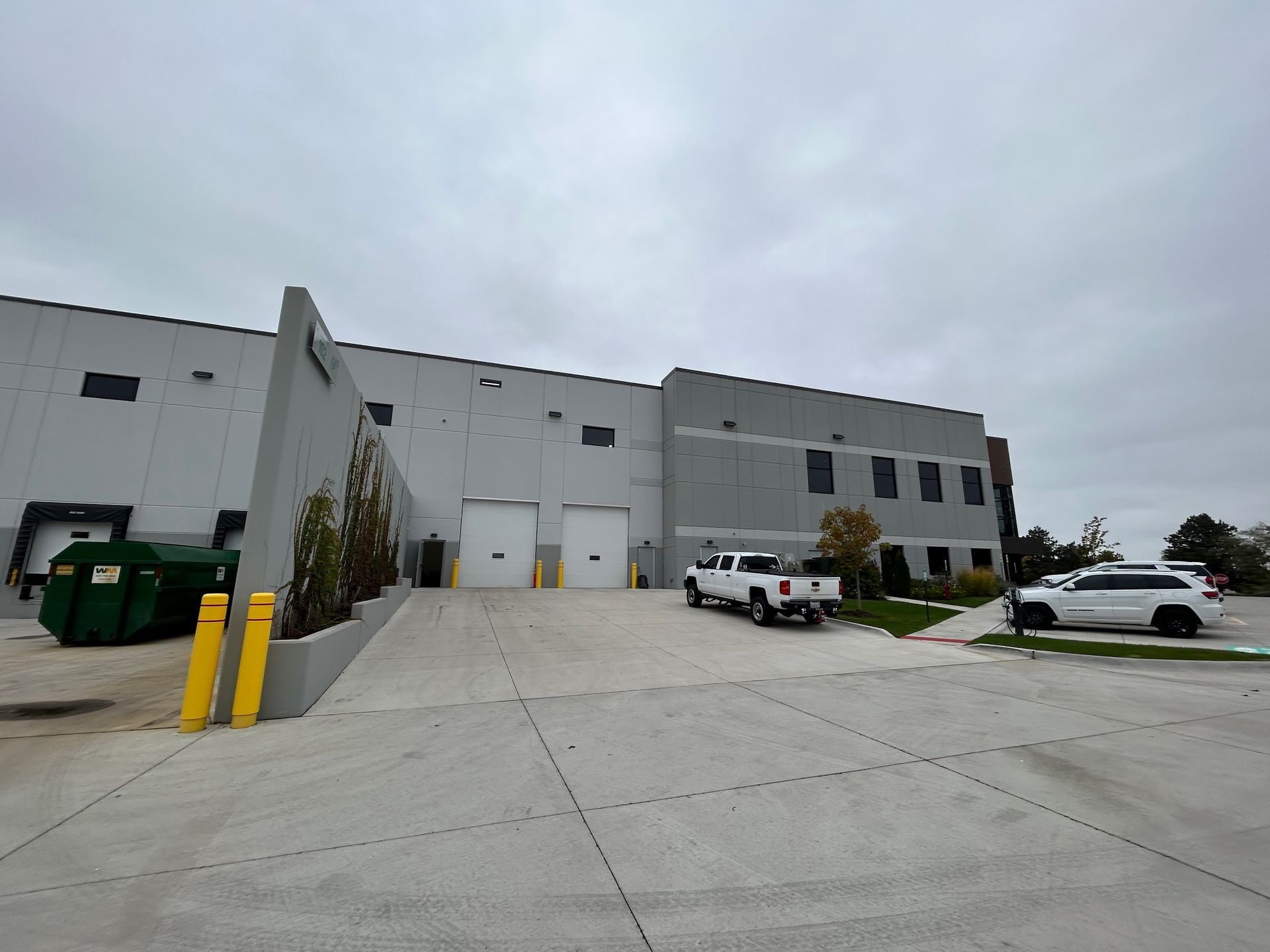 Image of a large warehouse facility during a Soft Washing Commercial Exterior Cleaning service, showing freshly cleaned concrete surfaces, loading dock doors, parked vehicles, and industrial dumpsters outside the building.