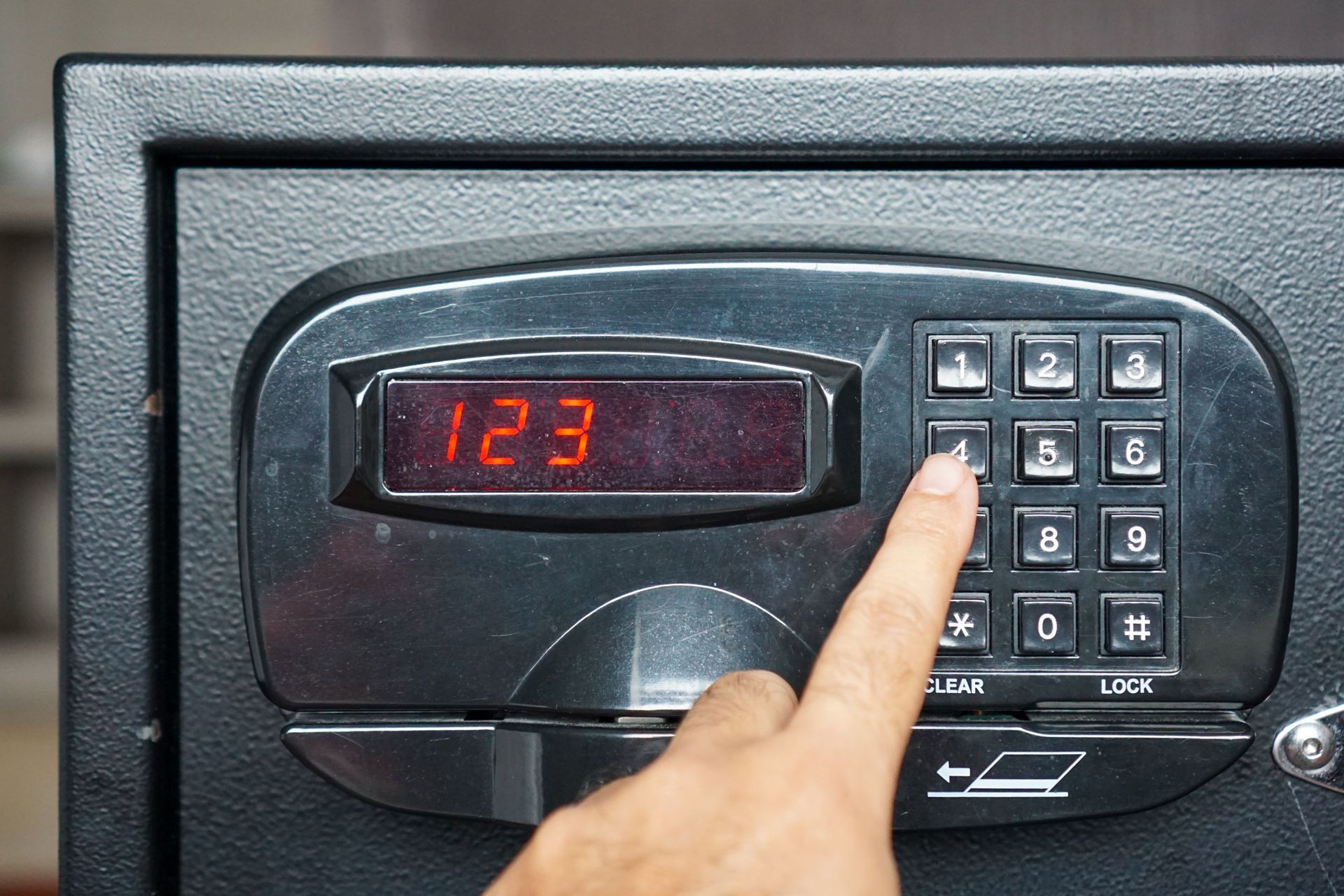 Finger entering code on a black electronic safe keypad with red display numbers.