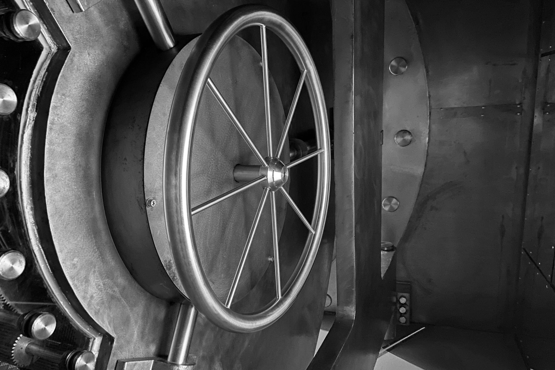 Black-and-white close-up of a large industrial metal wheel or vault door with spokes and bolts