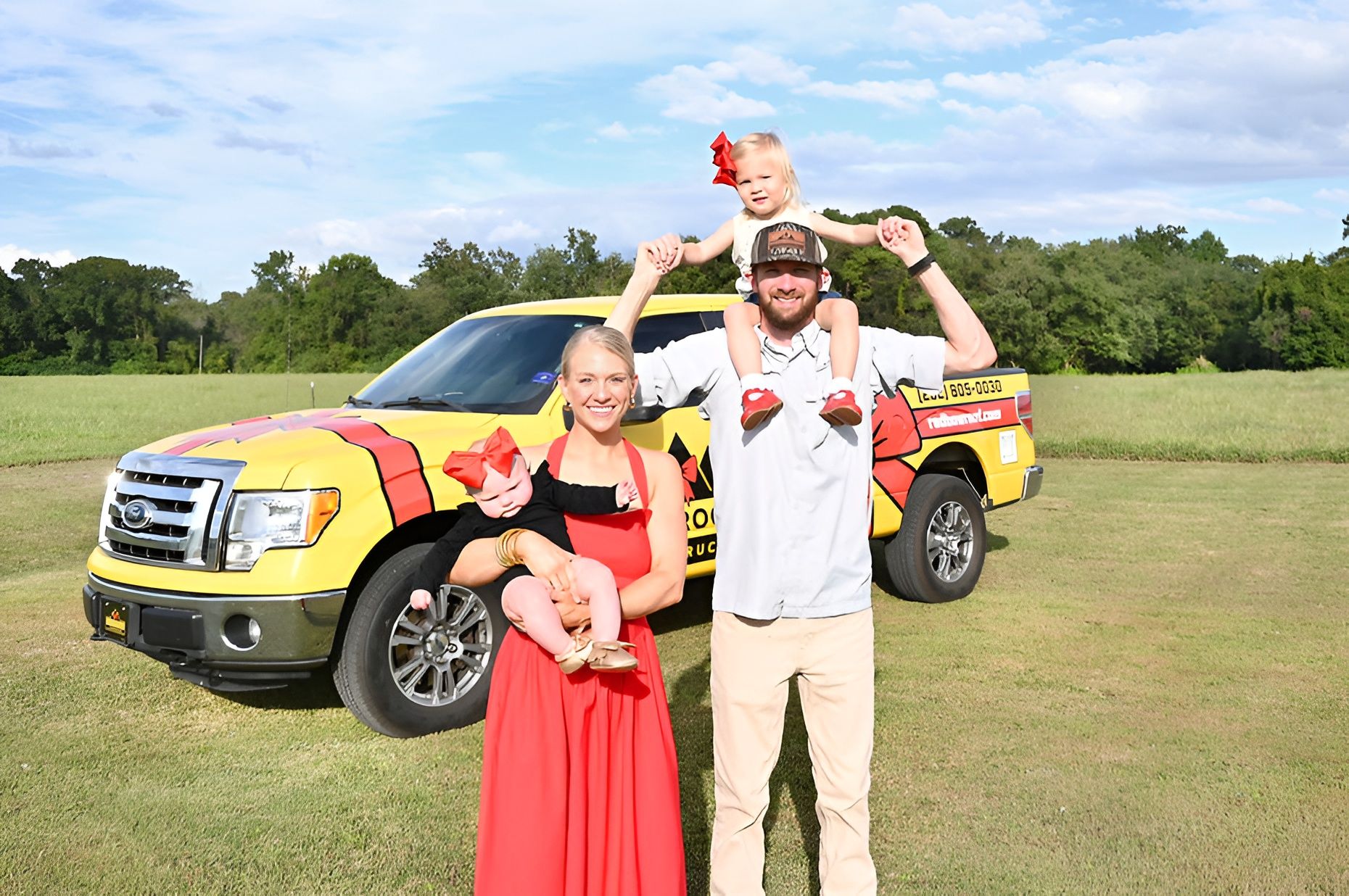 Four people posing by a yellow and red SUV in a grassy field, one seated on the hood and one holding a child.