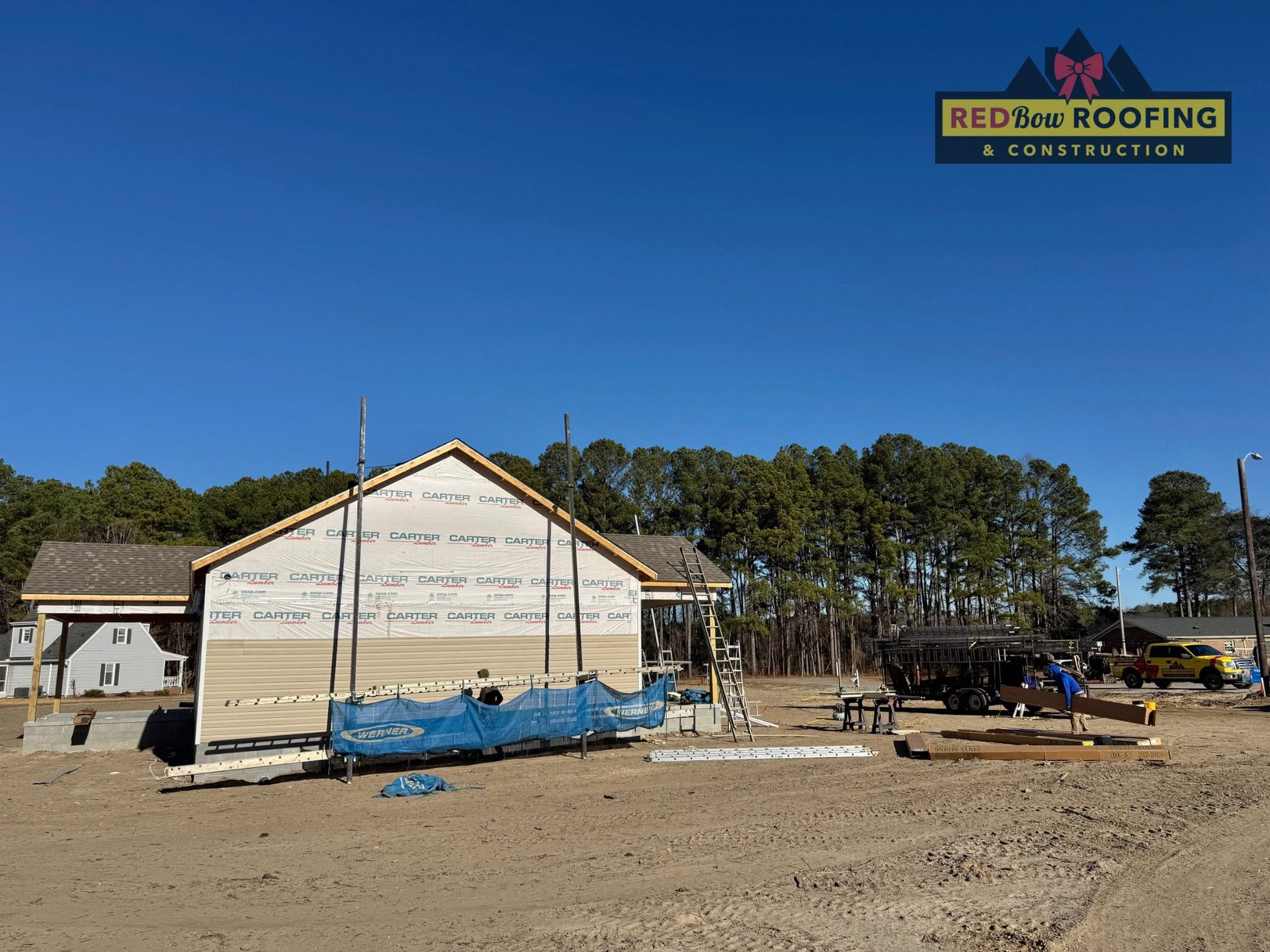 House under construction with wrap and blue tarp under a clear blue sky, construction logo in corner