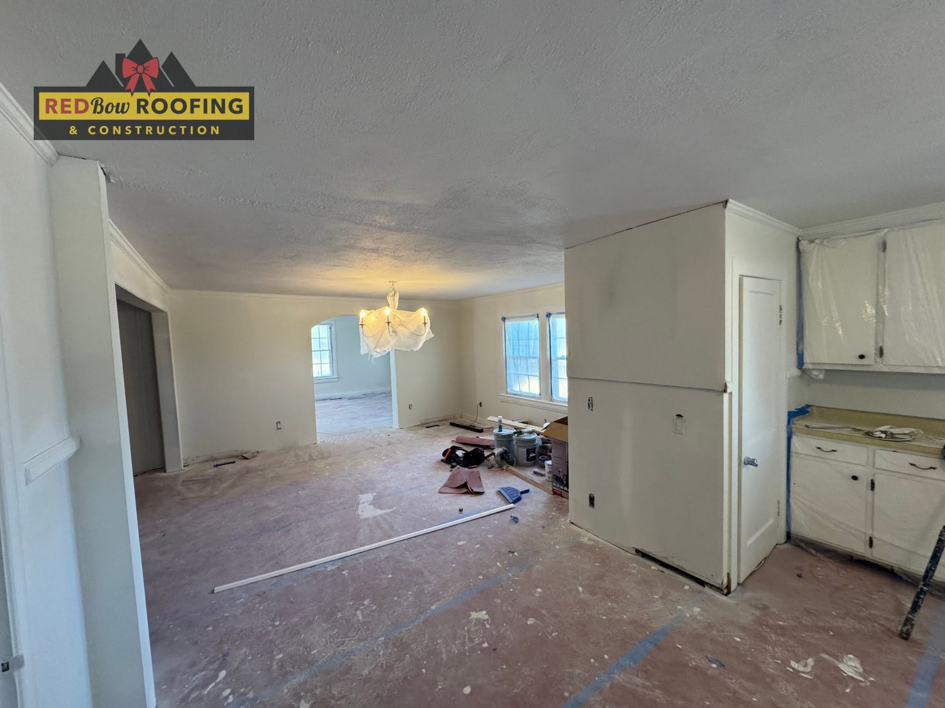 Damaged living room with debris on the floor, white walls, and open kitchen cabinets on the right.
