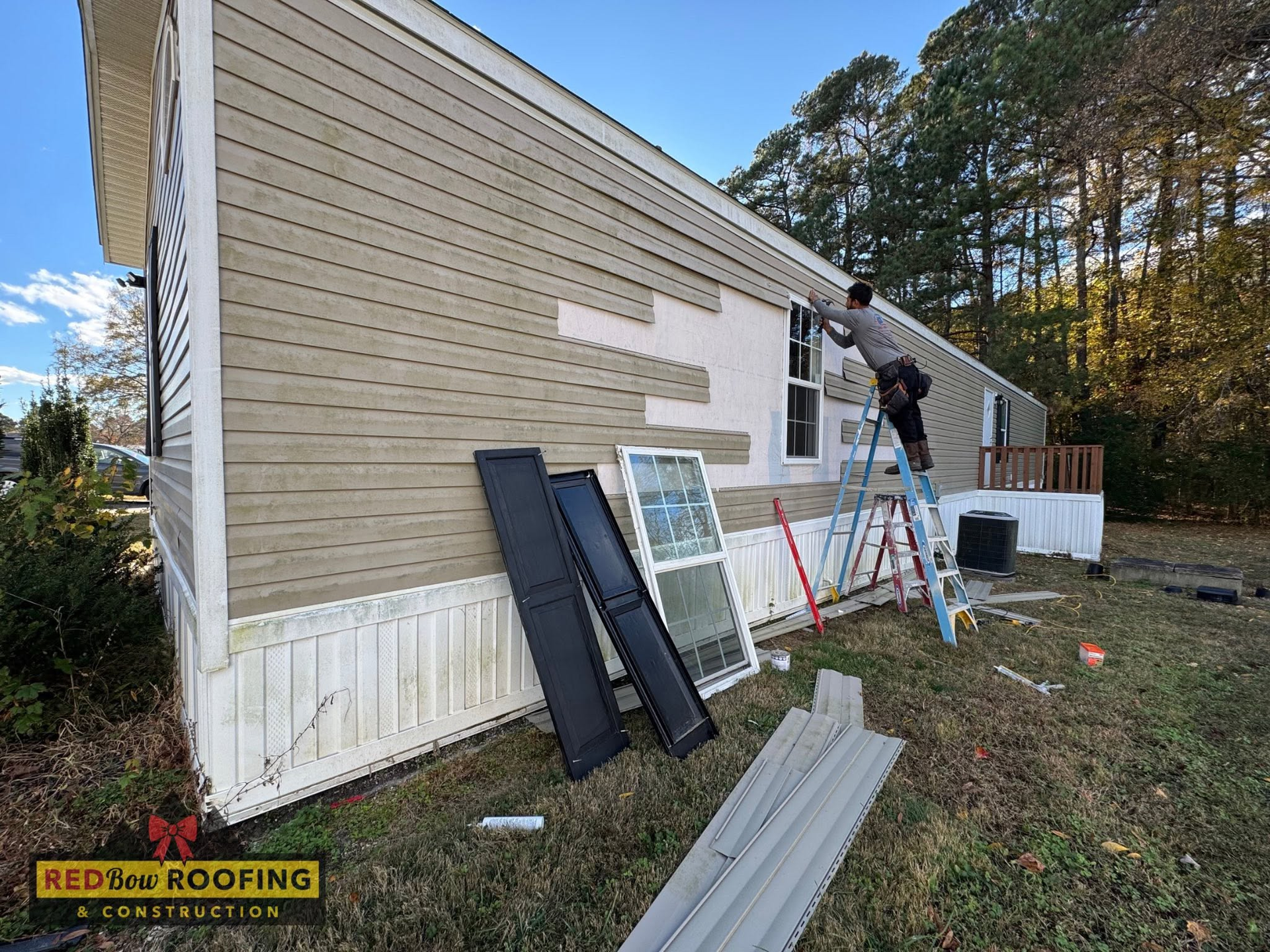 Worker on ladder repairing beige house siding with roofing materials stacked outside.