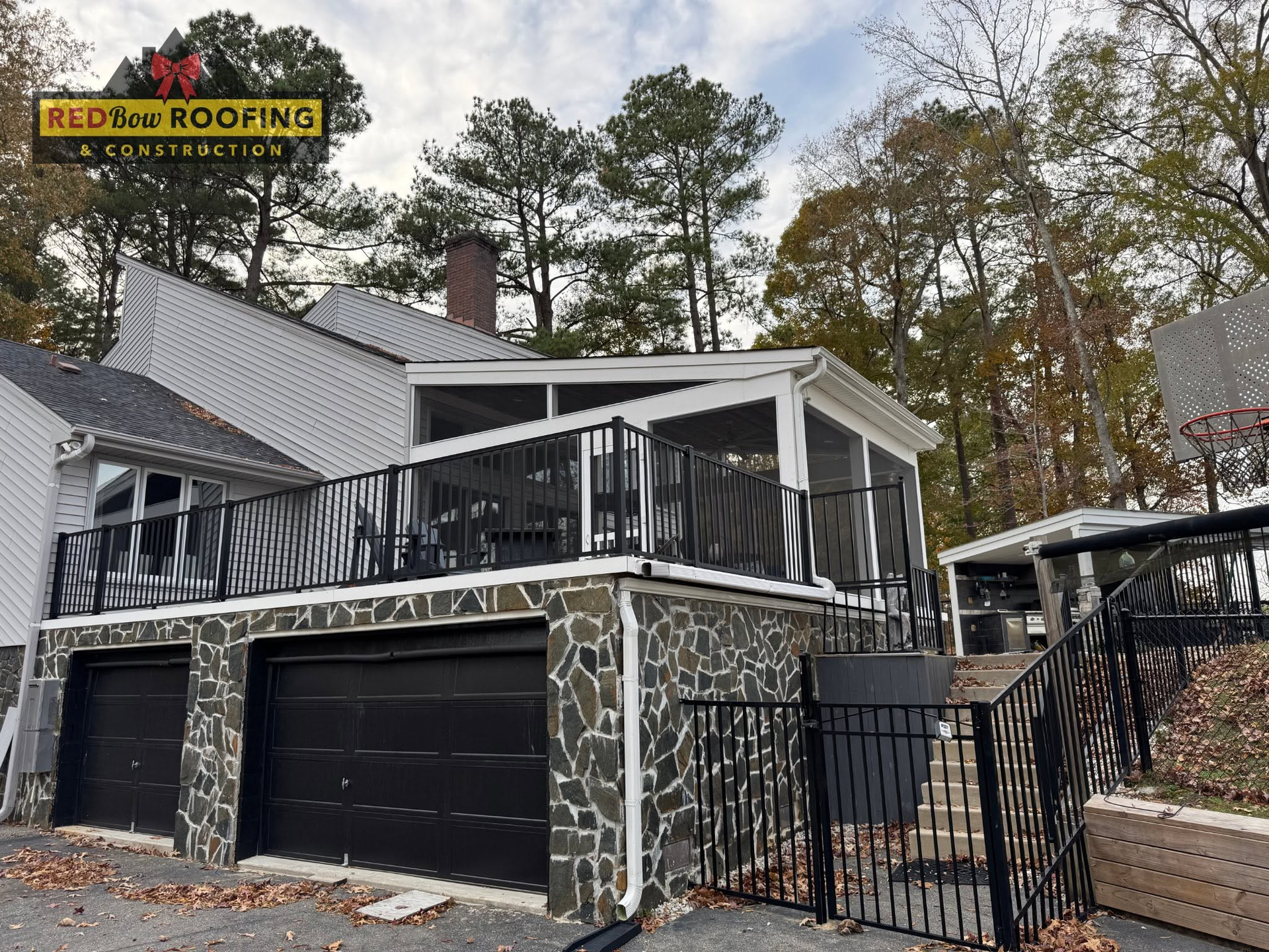 Stone-faced house with black garage doors, balcony, and fenced front yard under a cloudy sky