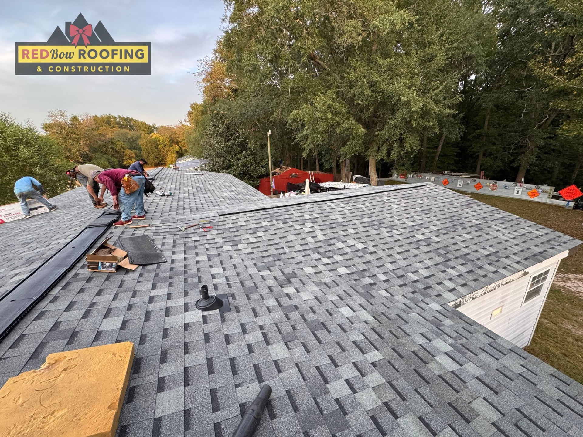 Roofers installing gray shingles on a large house roof, with trees and a logo in the corner.