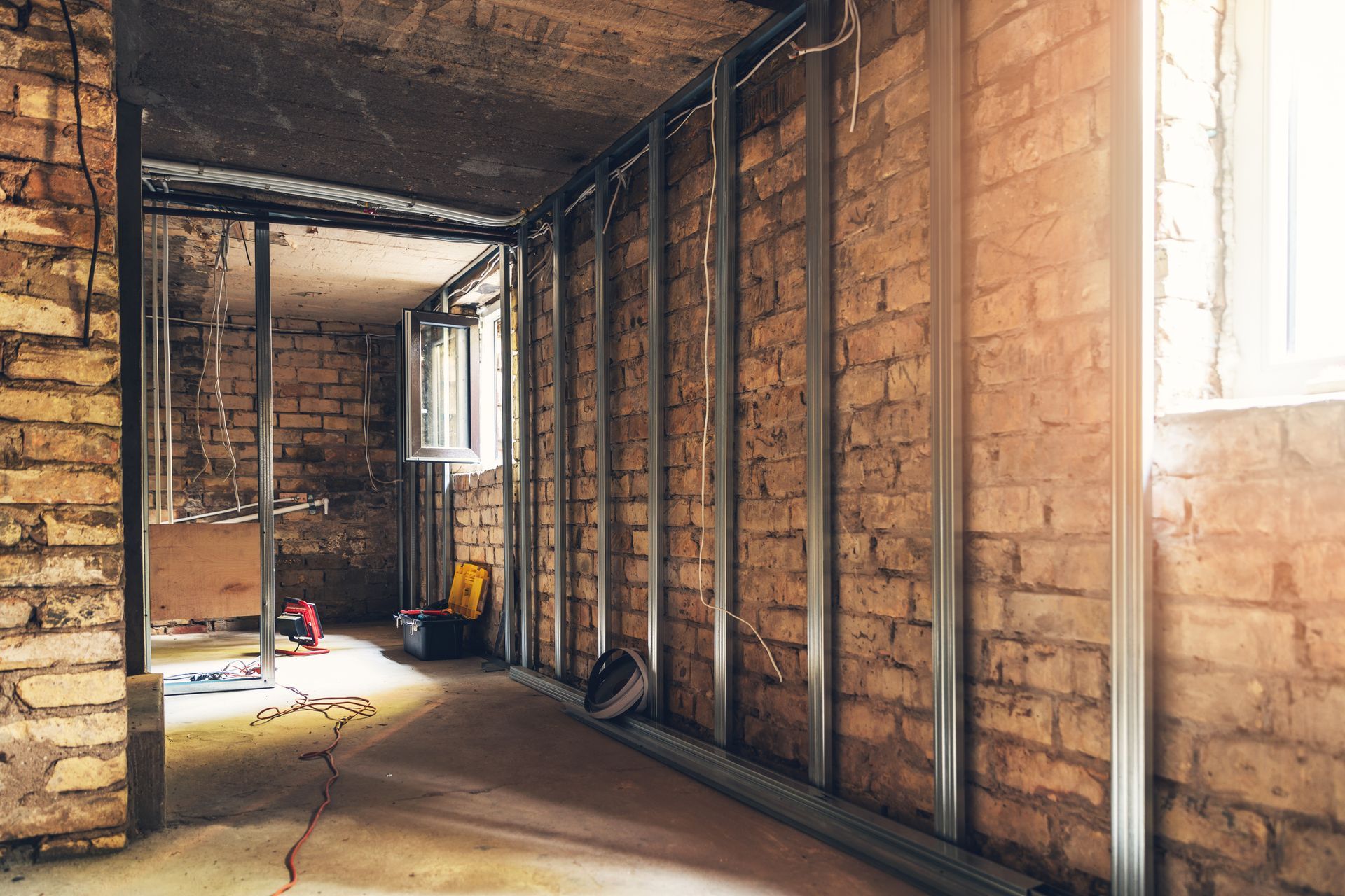 Unfinished brick room with exposed studs, sunlight through windows, and construction tools on the floor