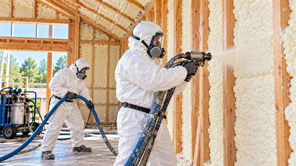 Two spray foam insulation technicians wearing full white protective suits, respirators, goggles, and gloves while installing spray foam insulation in a residential property in Tyler, Texas.