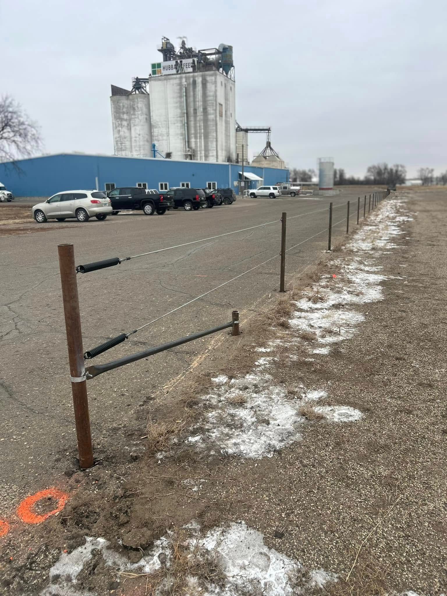 A barbed-wire fence runs along a gravel lot in front of industrial buildings and parked vehicles on a cloudy day.