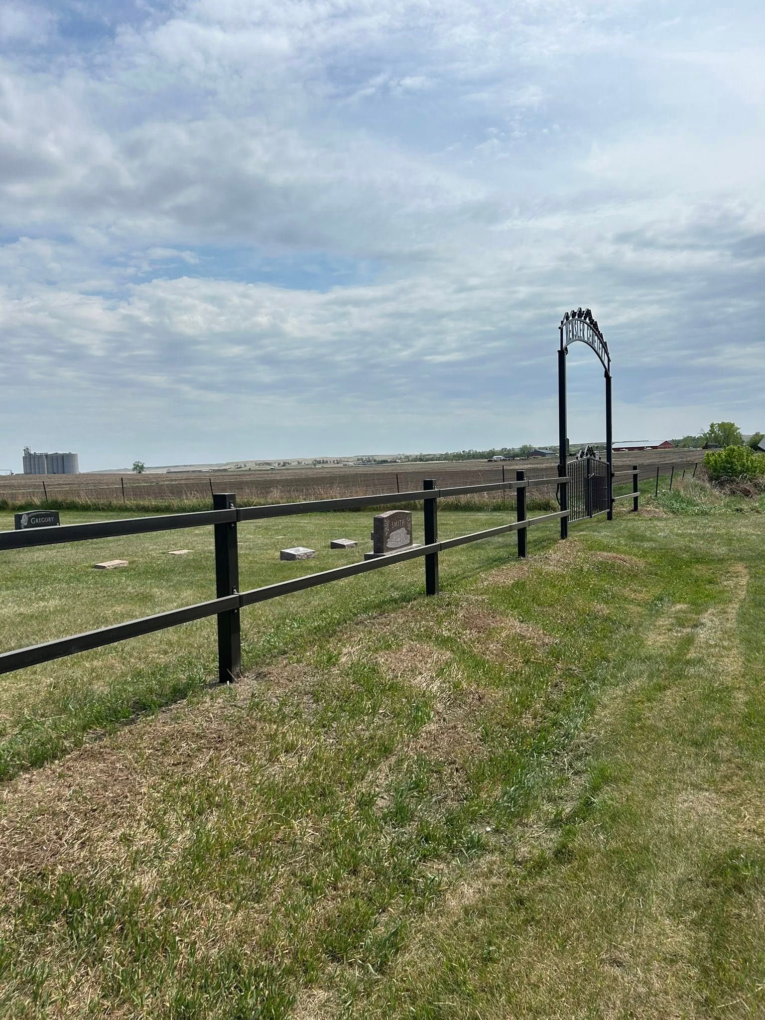 A cemetery with a black fence, gate, and tombstones on a grassy field under a cloudy sky.