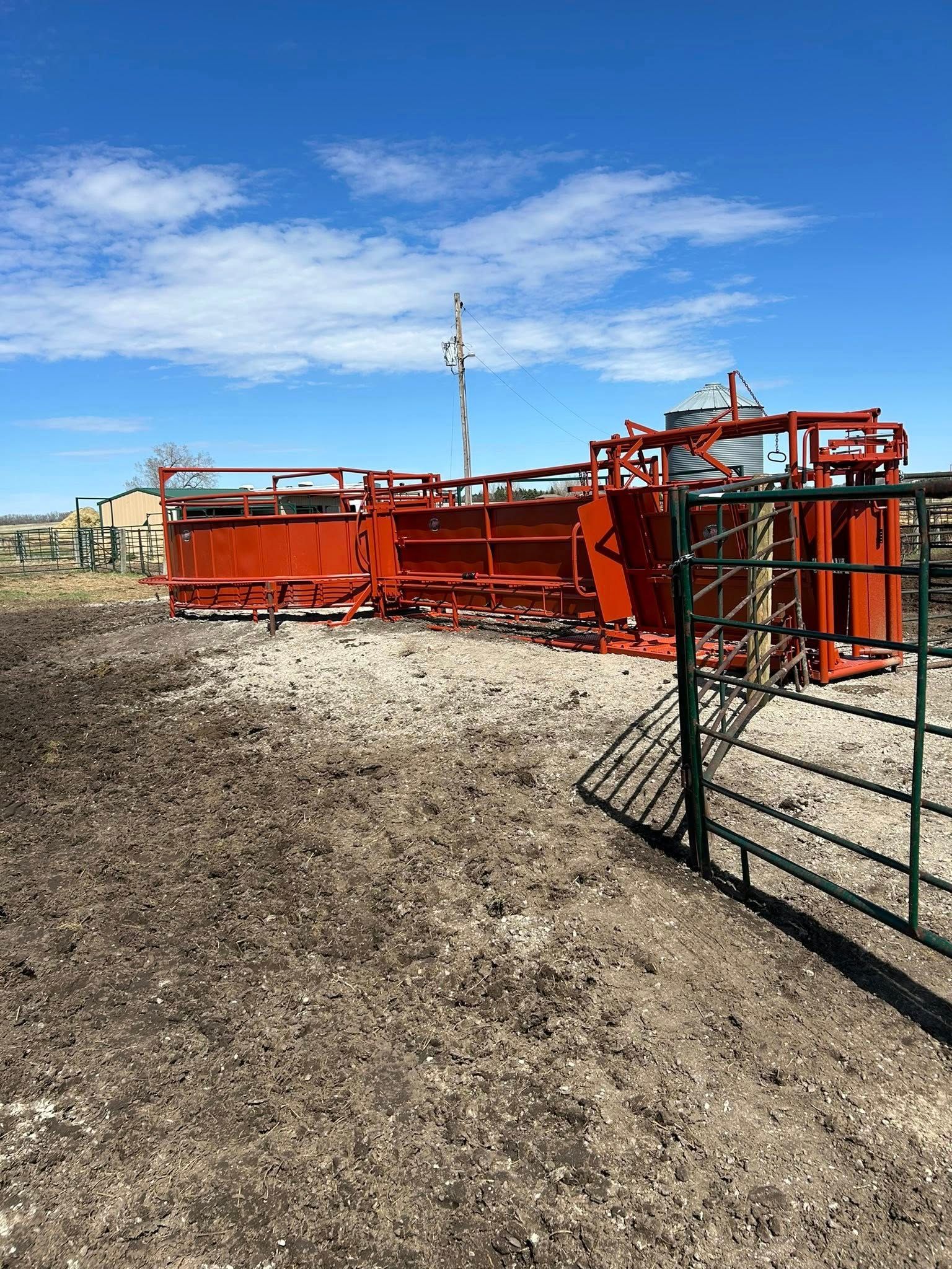 Red metal cattle chute and pen on a dirt ground under a blue sky.