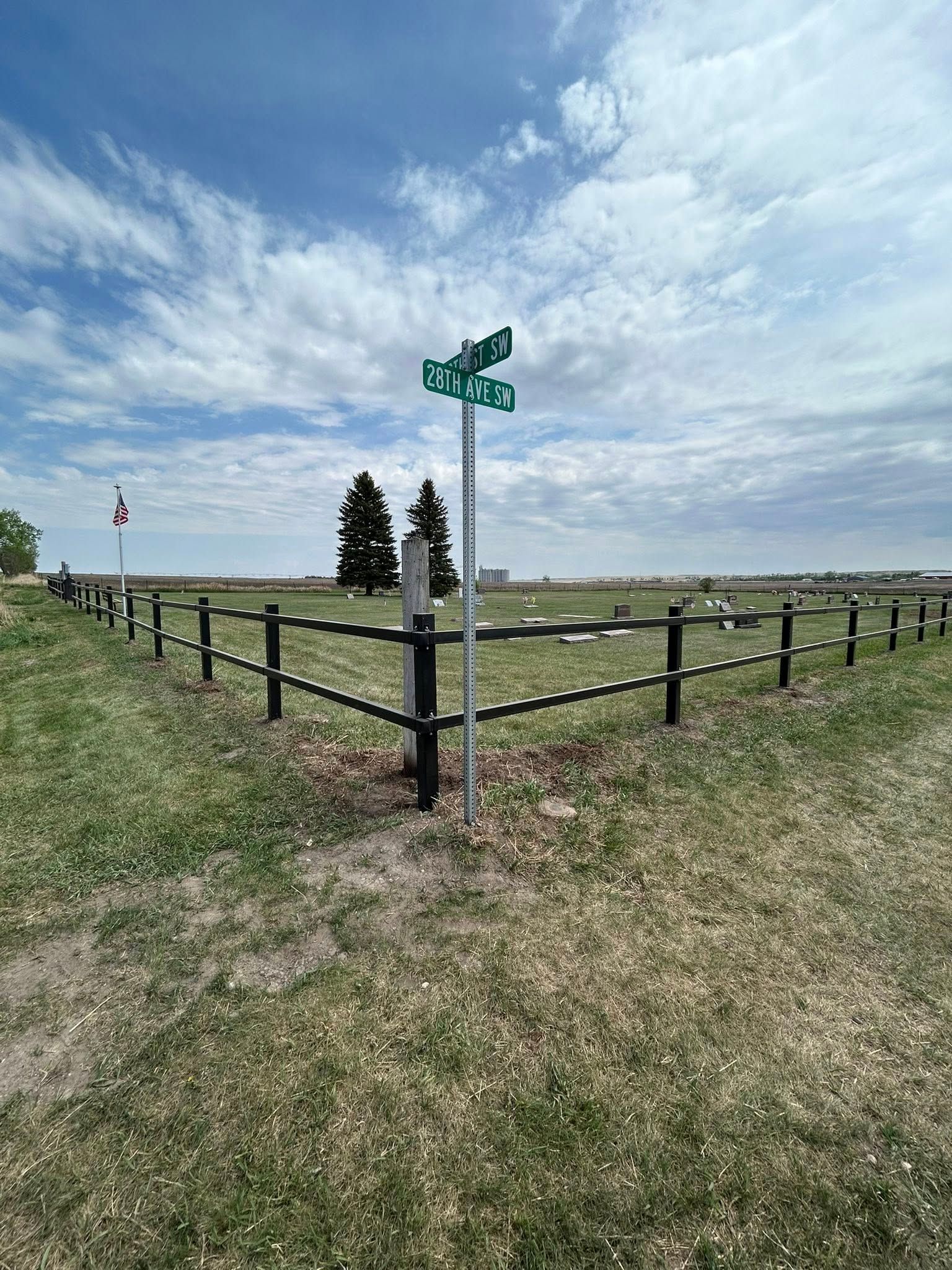 Signpost at a cemetery corner, framed by a black fence, against a cloudy blue sky.