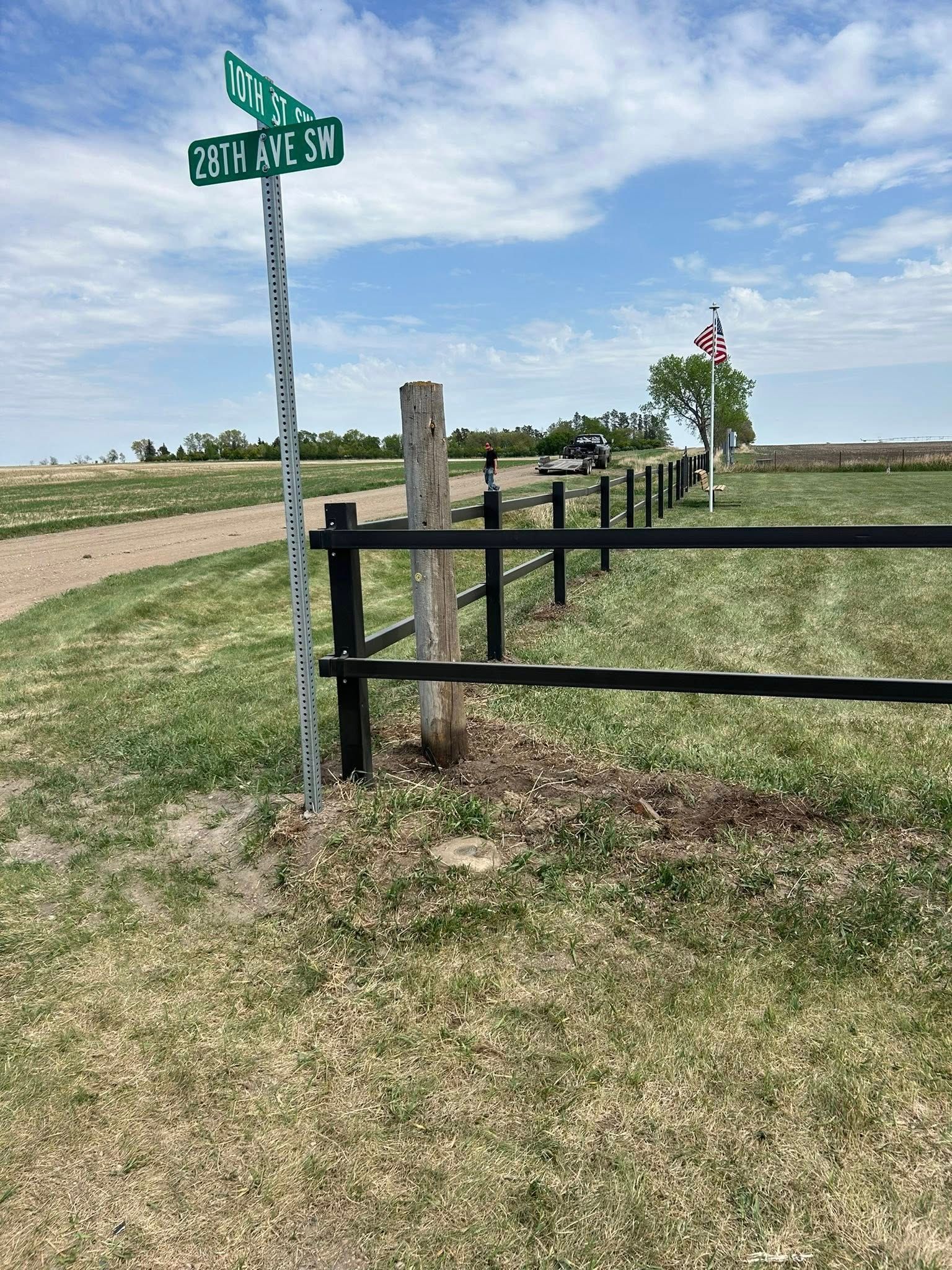 Green street sign at a corner with a black fence and a flag against a blue sky.