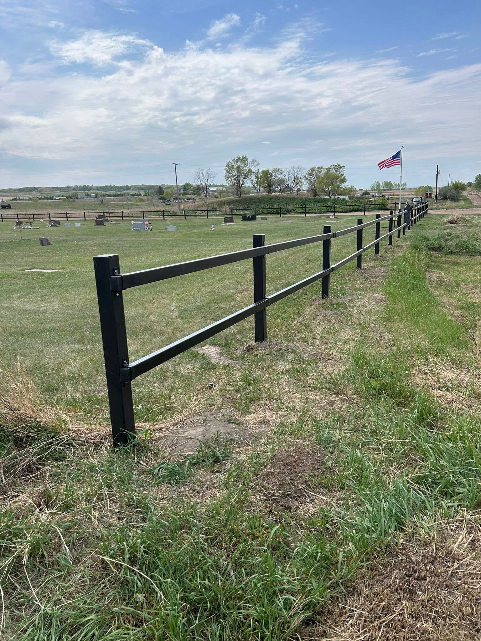Black fence in a grassy field, cemetery in the background, American flag waving.