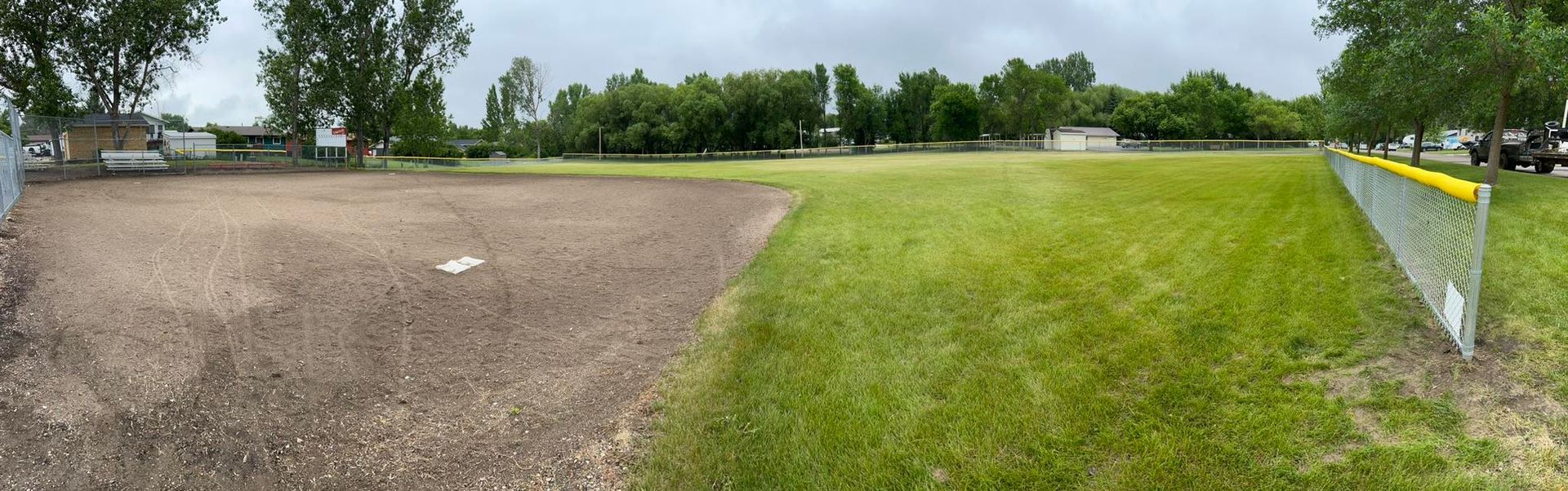 Baseball field, half dirt infield, half grass outfield. A gray sky. Trees in the background. Chain-link fence on the right.