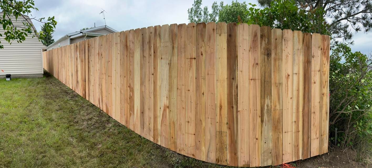 A wooden fence curves around a grassy yard, near a house and trees. Overcast sky.