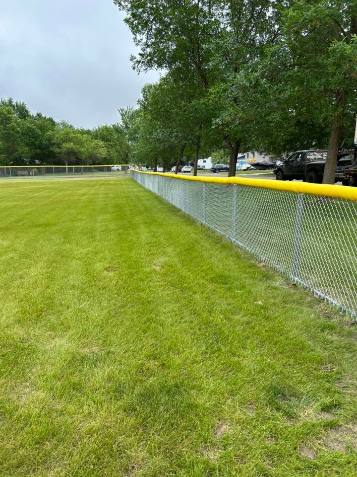 Chain-link fence with yellow top rail along a grassy field, trees in background, overcast sky.