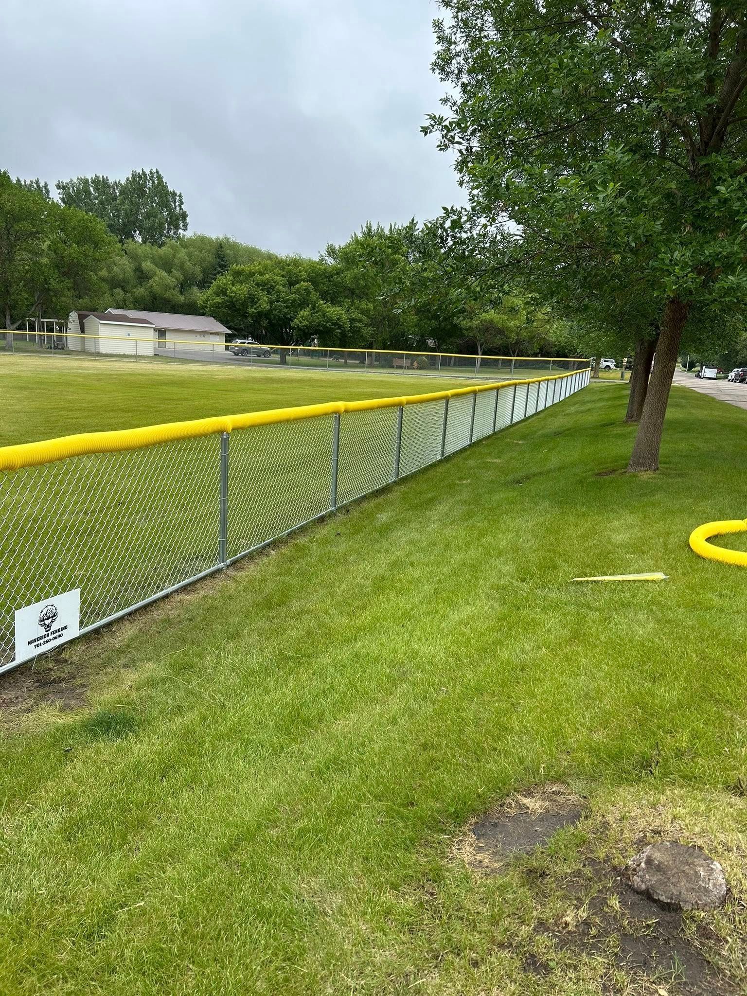 A chain-link fence topped with yellow padding lines a grassy area, trees and buildings in the background.
