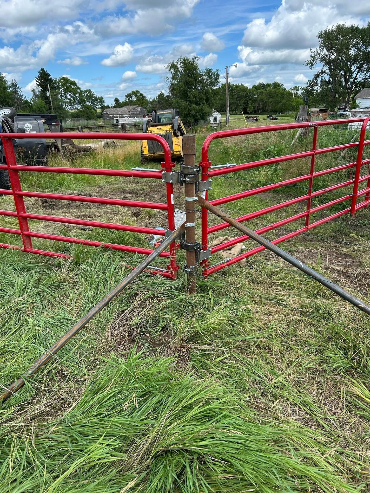 Red metal gate, open, blocking a grassy field. A yellow excavator is visible in the distance.
