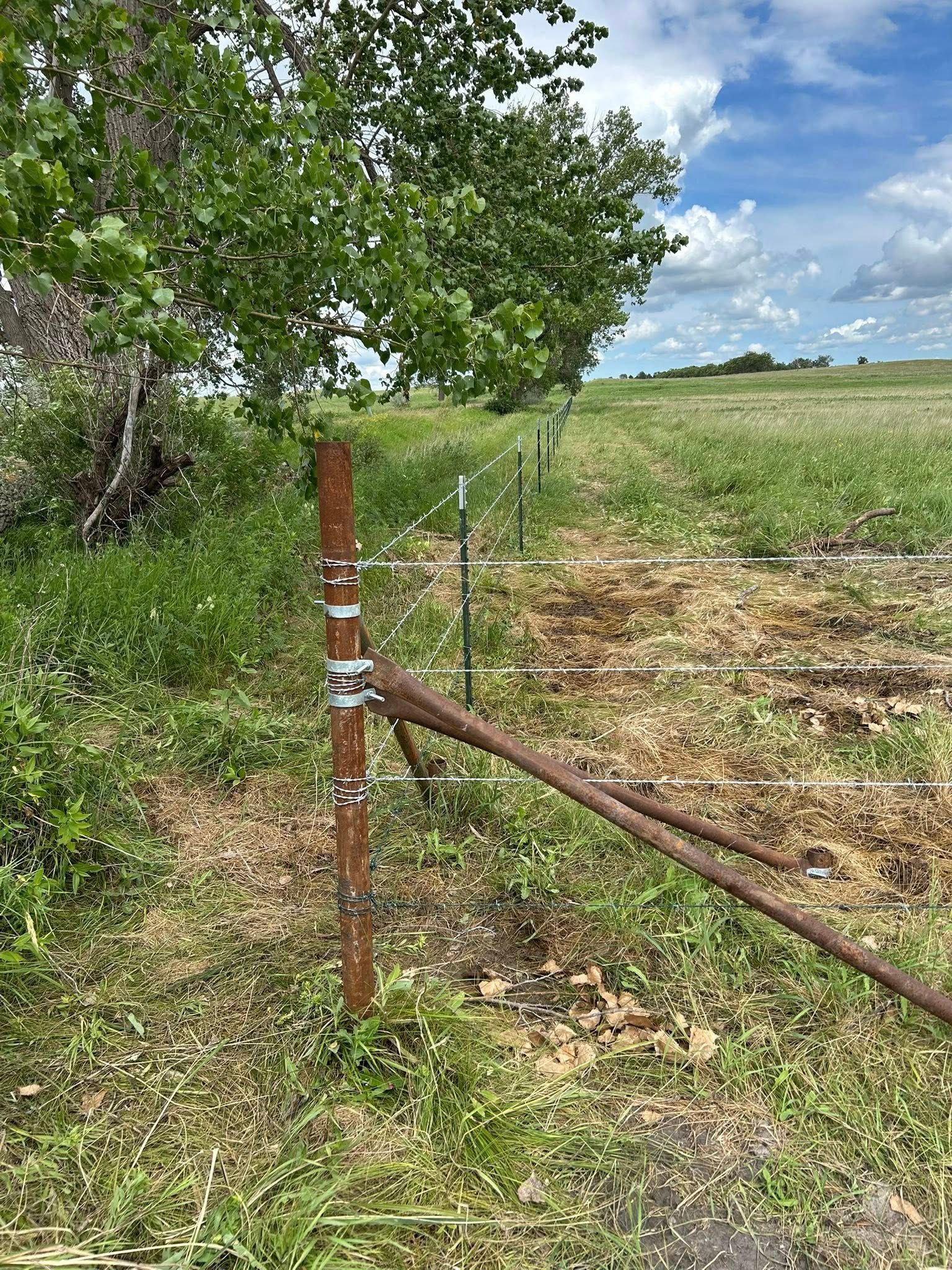 Rusty metal fence post with wire strands and a metal lever in a grassy field.