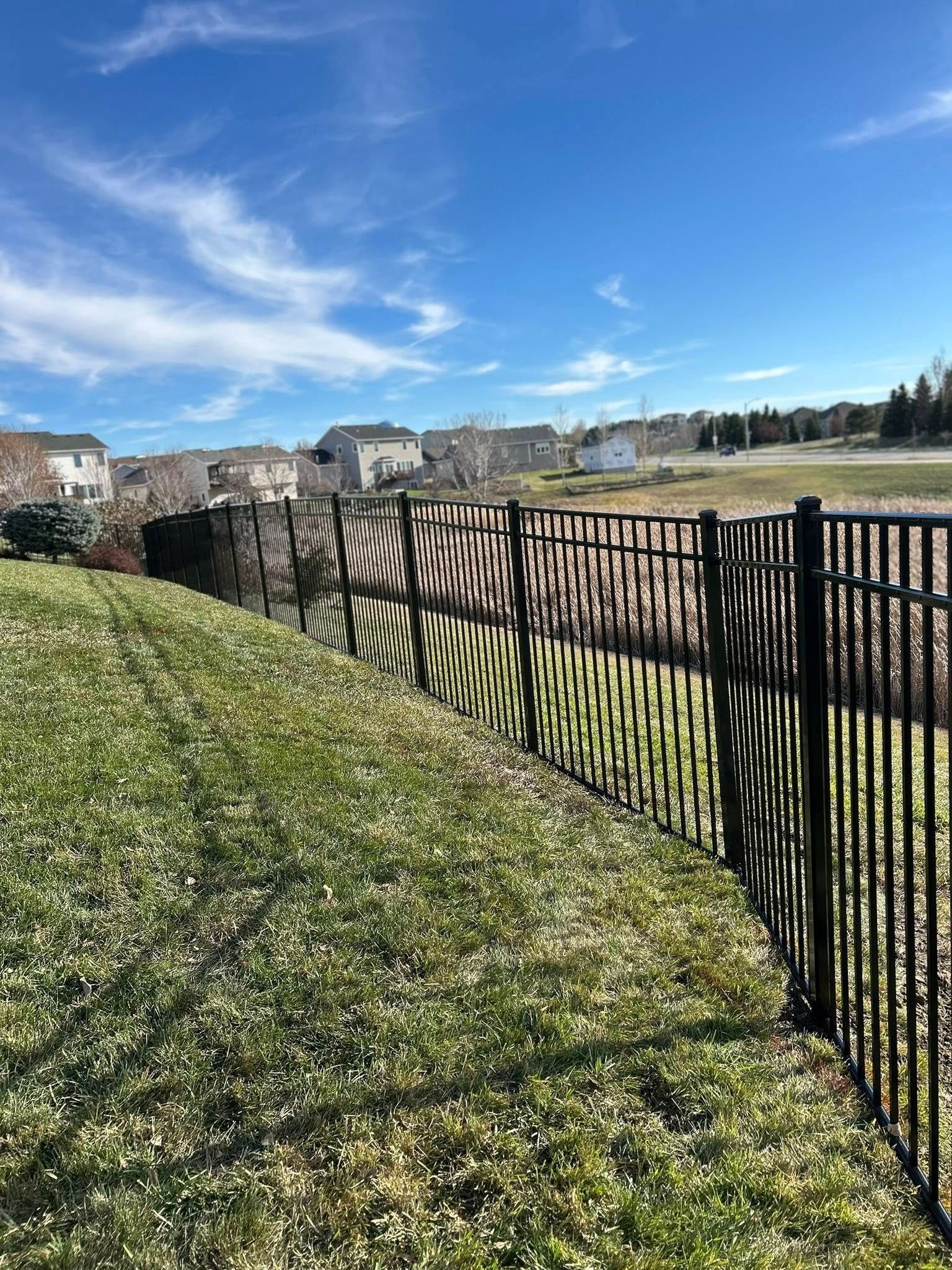 A black metal fence casts shadows on a lawn. Houses and blue sky with clouds in the background.